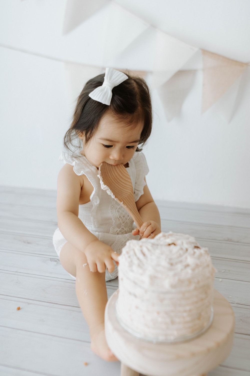 A small child in white outfit reaches toward a white frosted cake under party bunting.