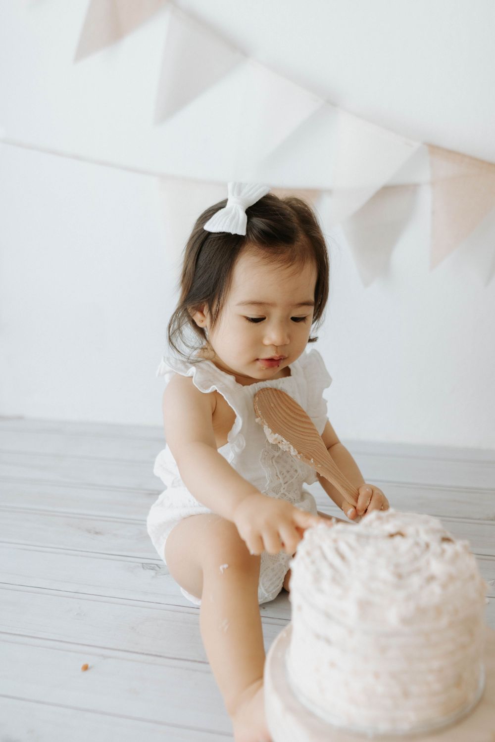 A toddler in lace romper sits beside a white cake on wooden pedestal with party decorations.