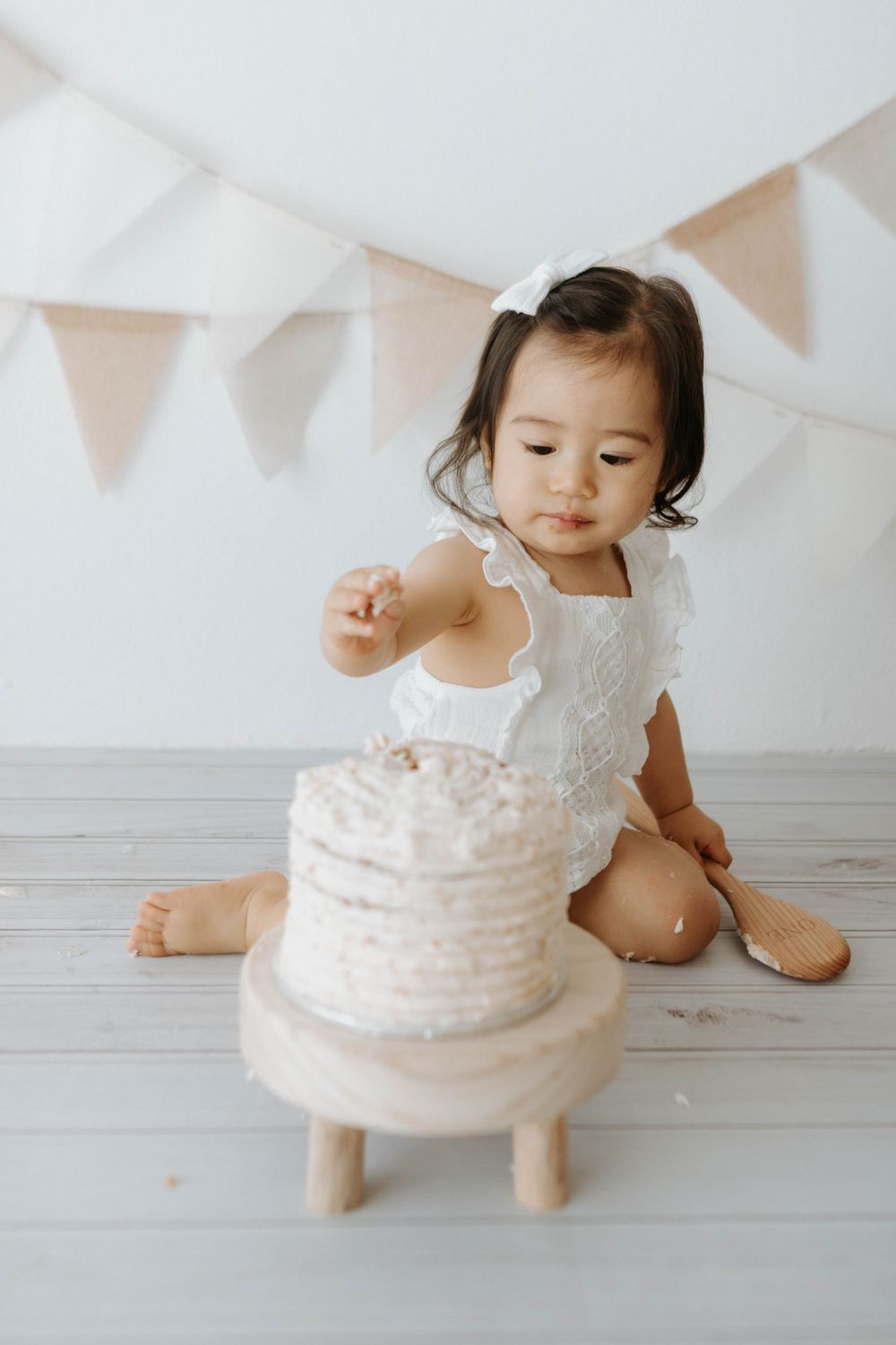 A small child in white outfit reaches for a white cake on wooden stand under party banners.