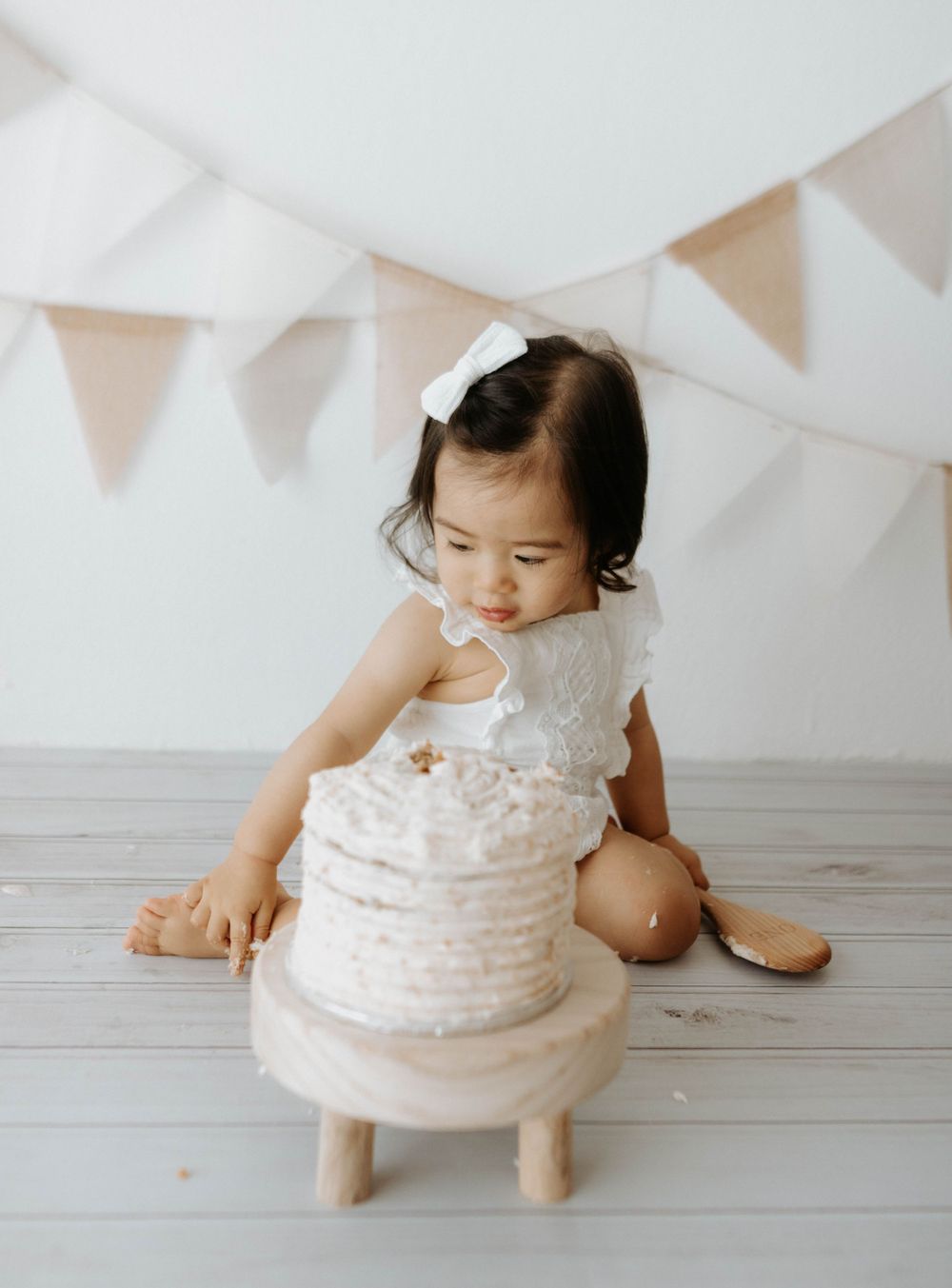 A toddler in white lace outfit sits beside a decorated white cake under festive bunting. cakesmash in a natural light studio in the mountain