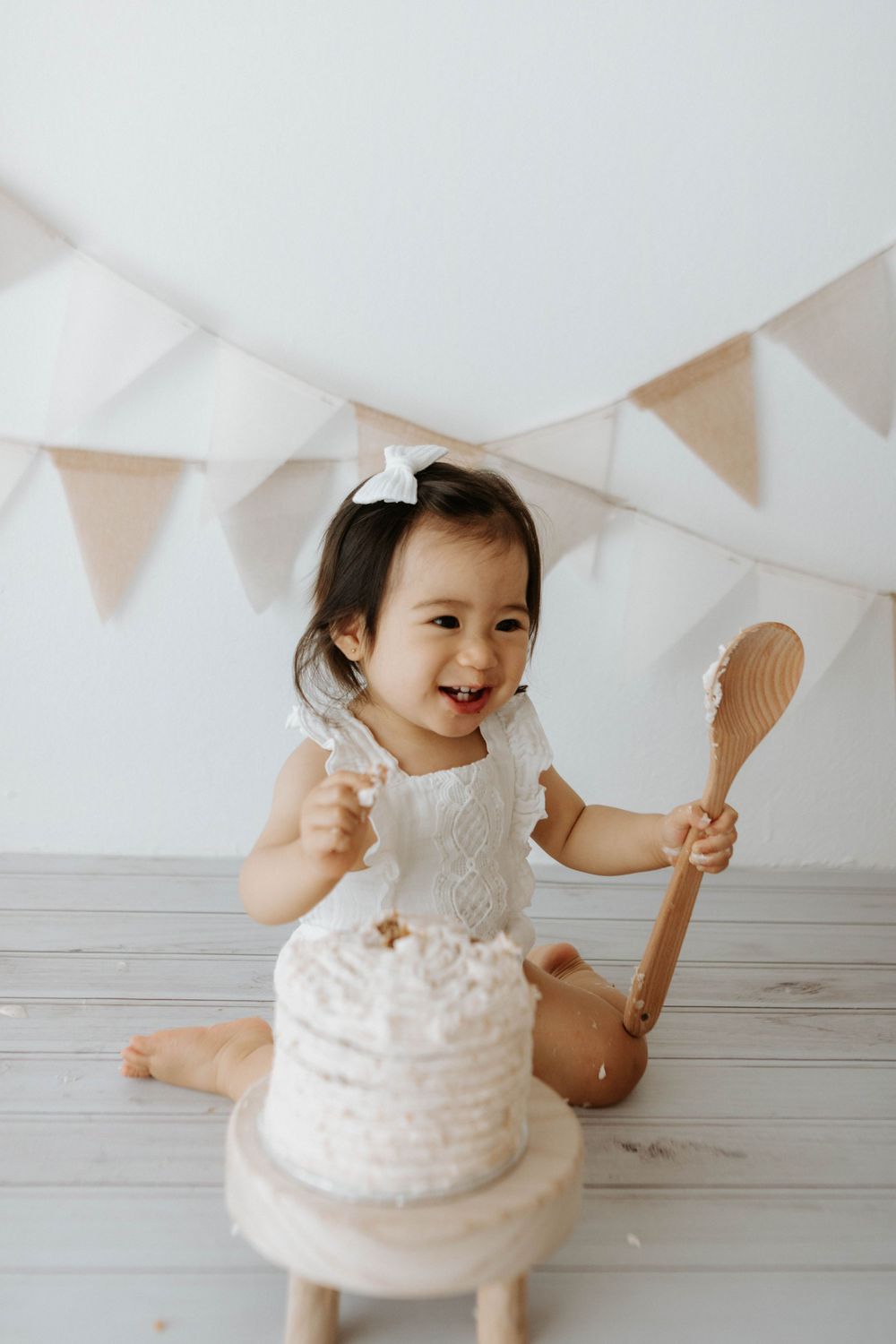 A small child joyfully plays with wooden spoons next to white cake under party bunting.