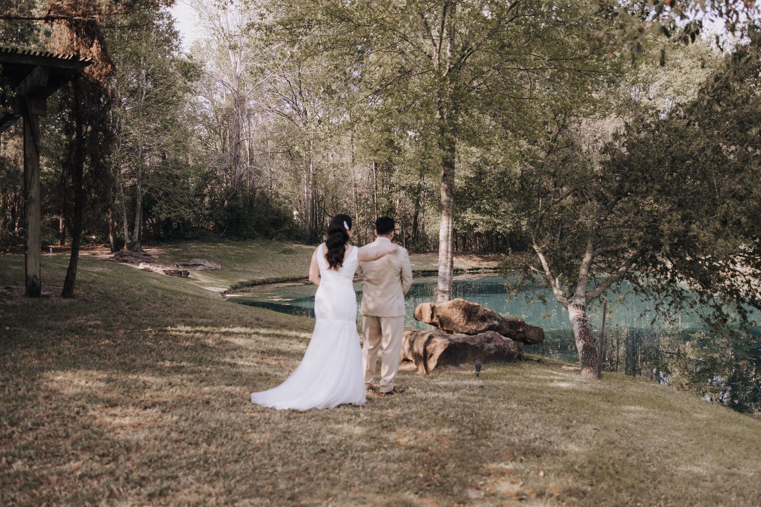 Wedding couple stands by a turquoise pond surrounded by trees in a dreamy outdoor setting.