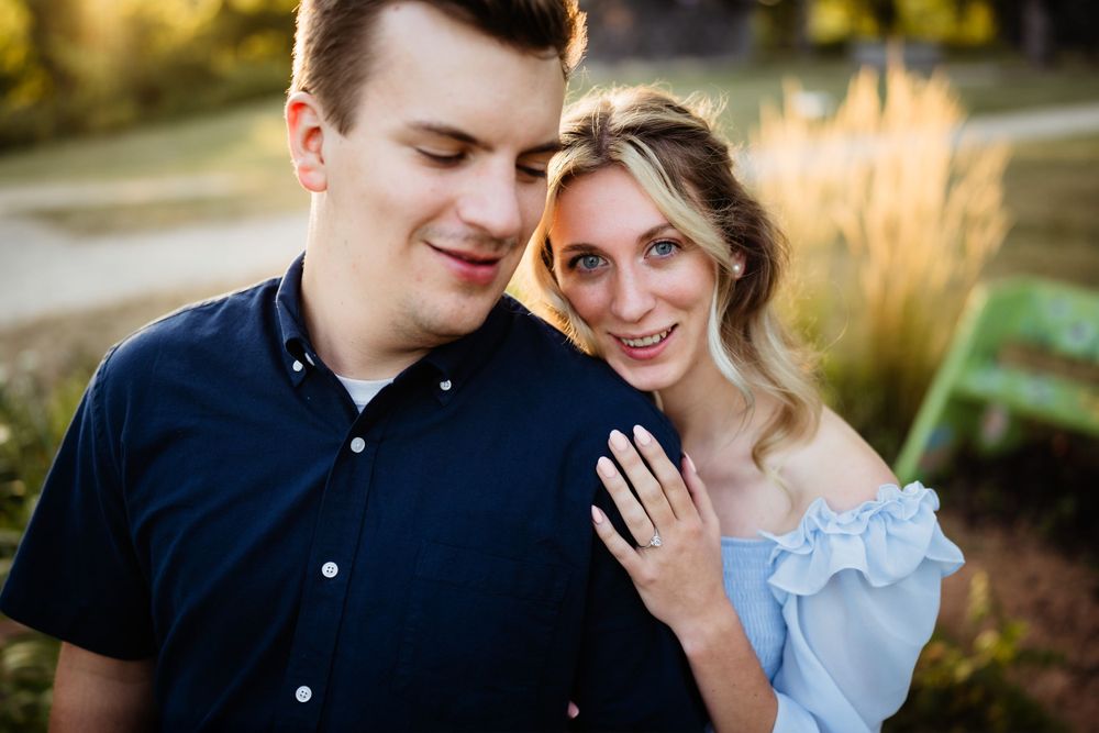 A couple in blue outfits poses together in a garden during golden hour while showing off an engagement ring.