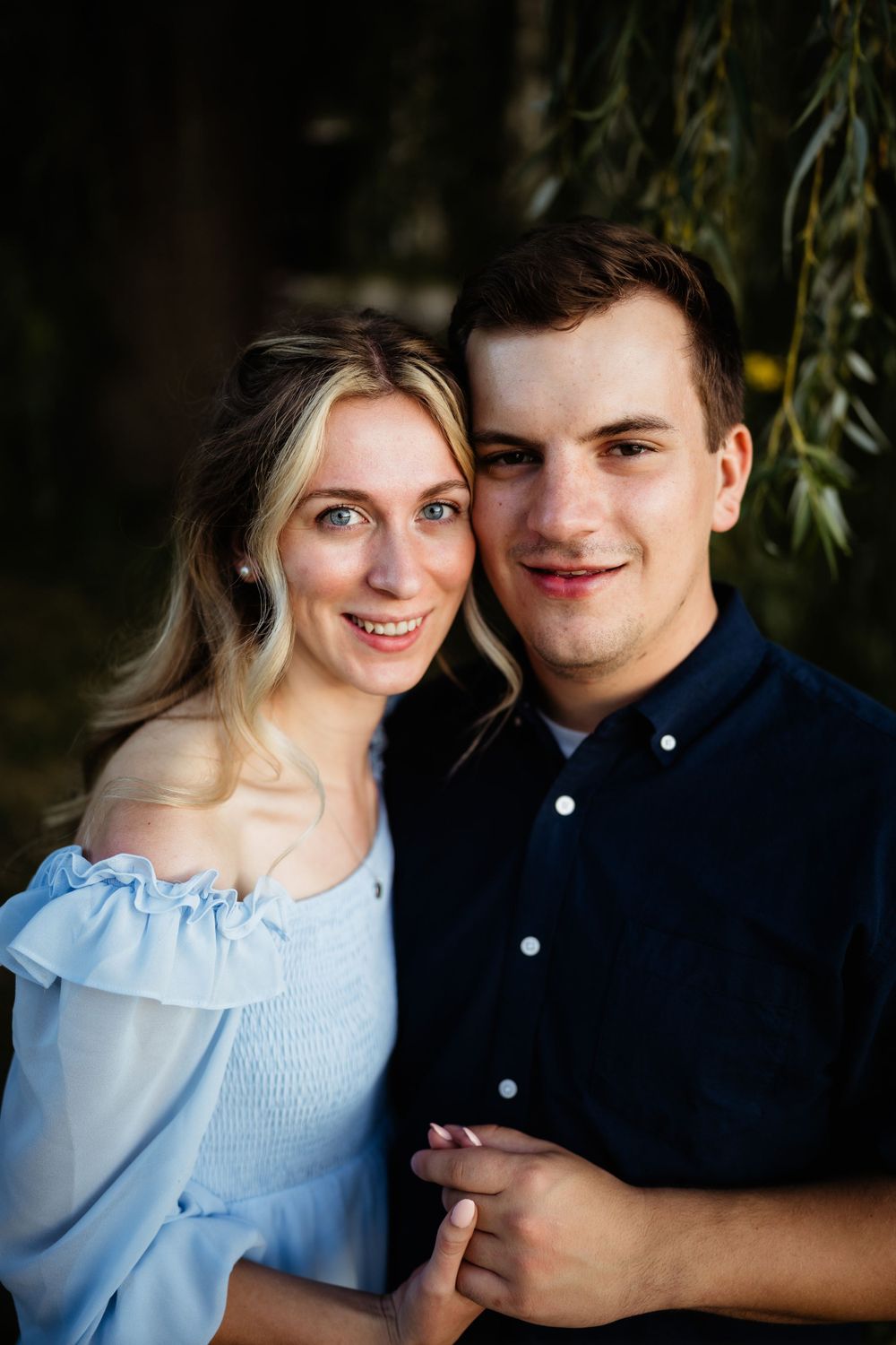 A couple in coordinating blue and navy outfits pose for a close portrait under willow branches.