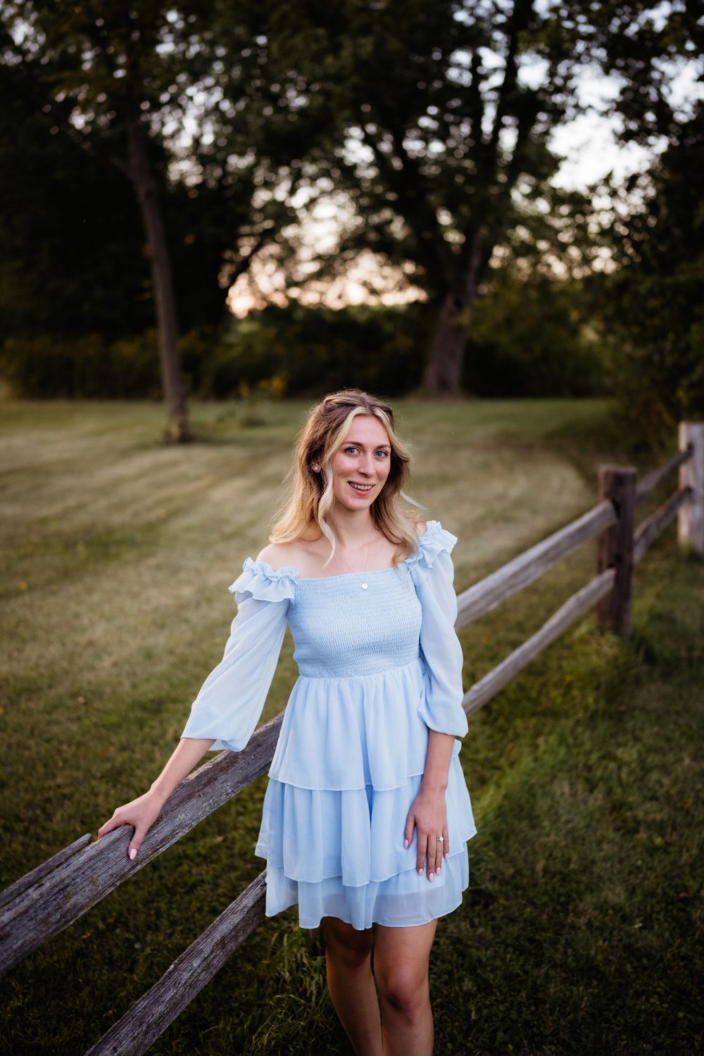 Woman in light blue dress posing by rustic wooden fence at sunset.