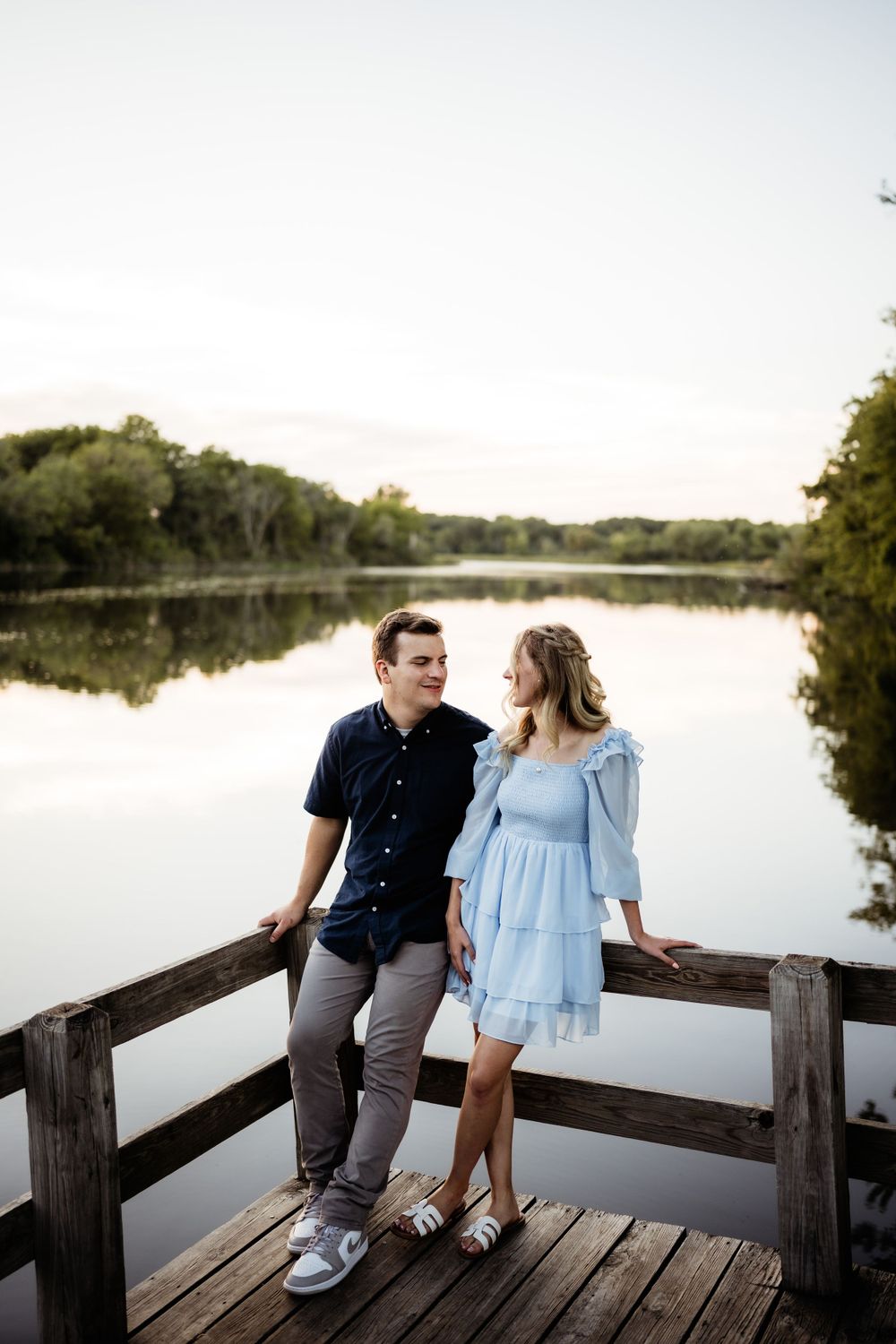 A couple in blue outfits sits together on a wooden dock overlooking a serene lake at sunset.