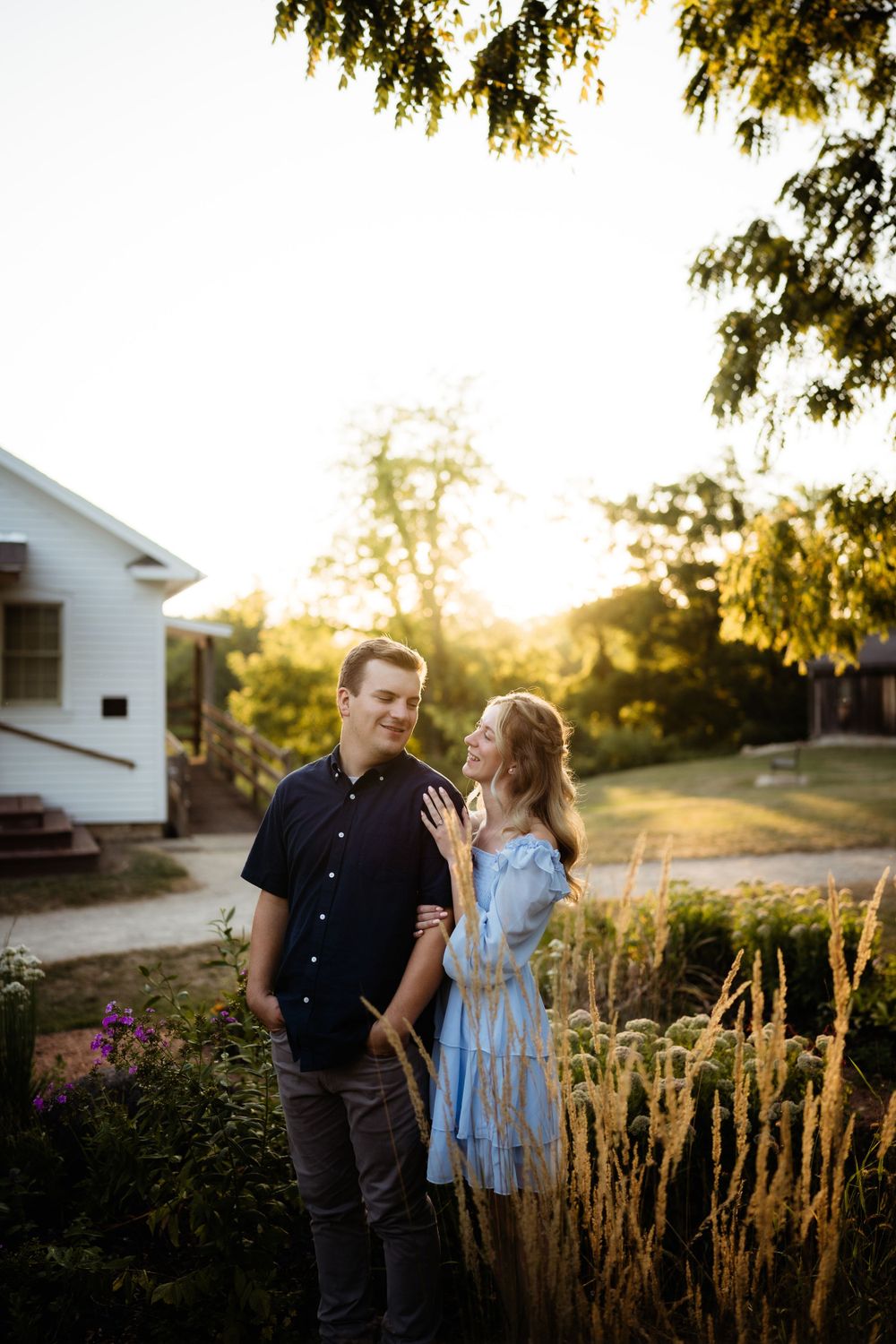 A couple shares a tender moment at sunset in front of a white house with ornamental grasses in the garden.