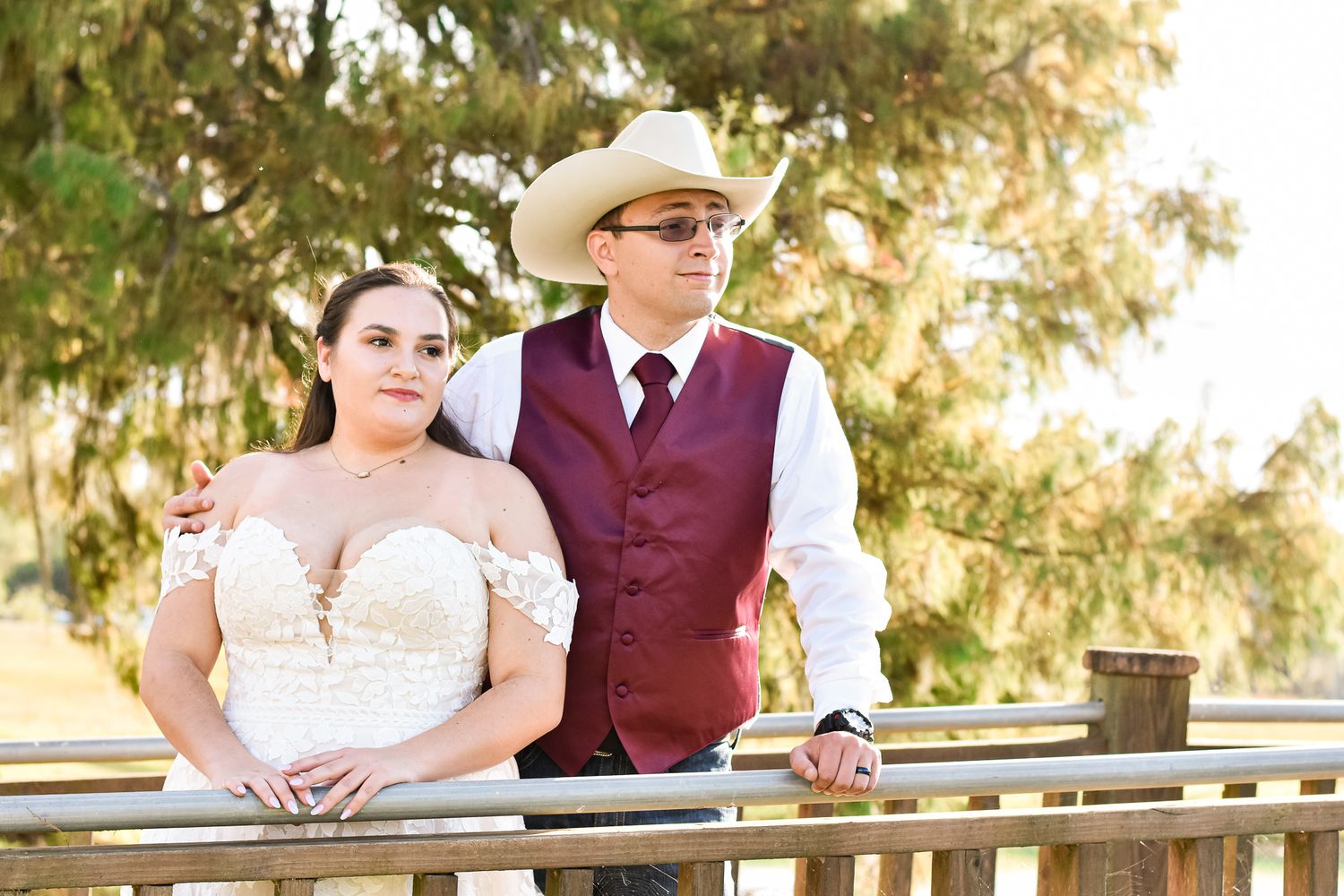 Romantic couple in formal attire pose against sunlit trees at outdoor venue.