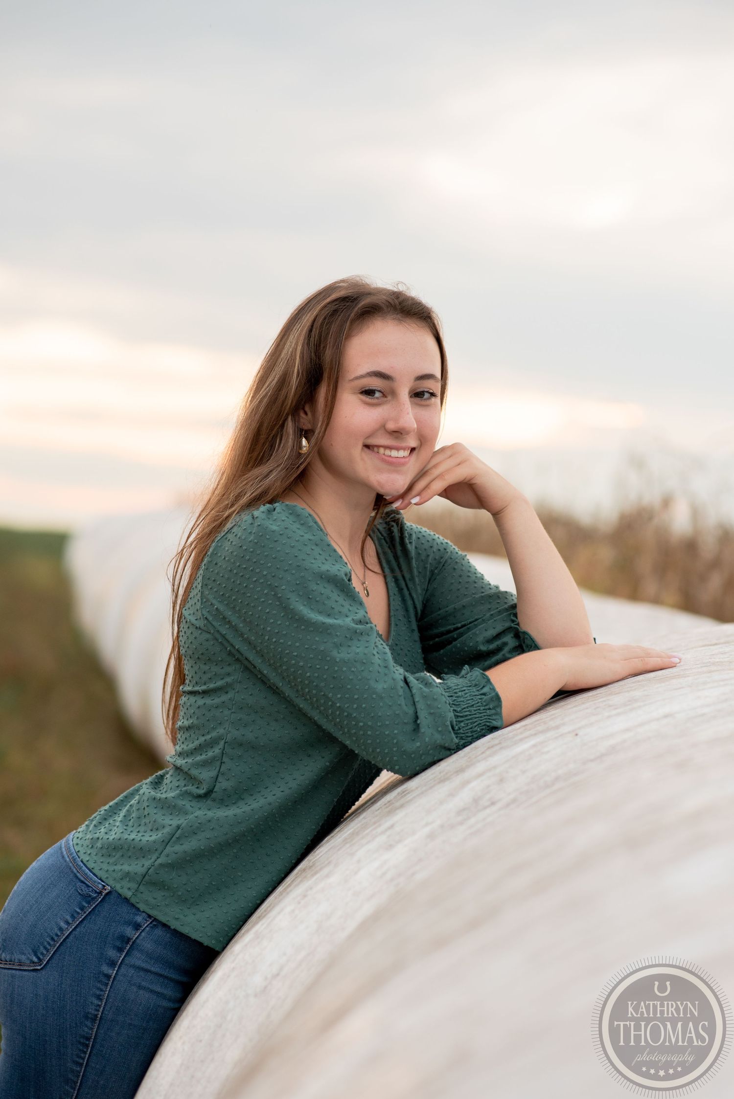 Casual outdoor portrait of someone in a green sweater leaning on a round bale of hay at sunset.