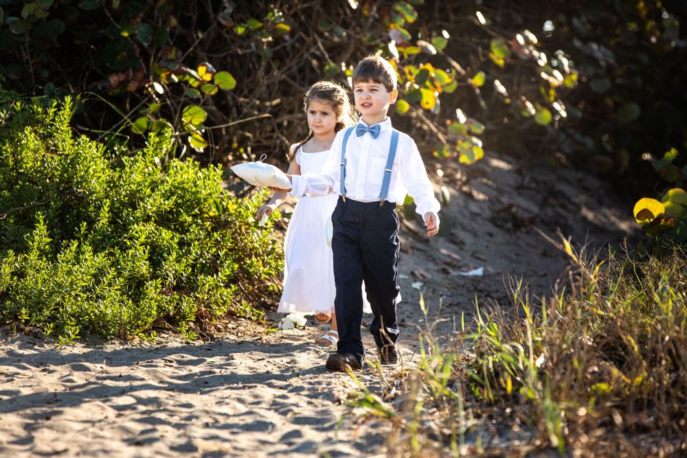 Two young children in formal attire walk together on a sandy beach path.