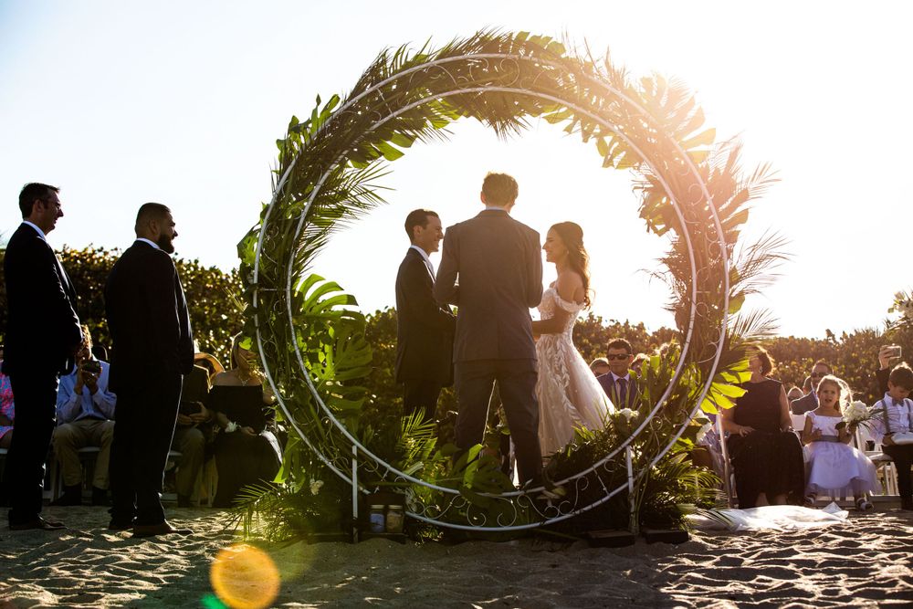 Sunlit wedding ceremony through a large circular floral arch with guests gathered around.