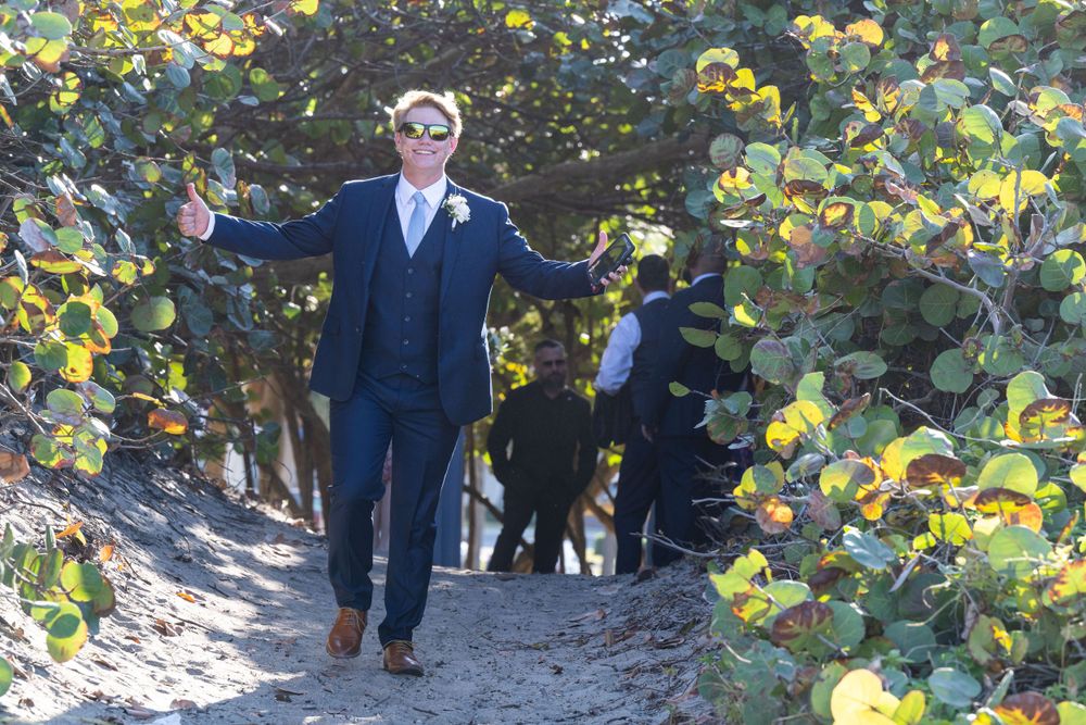A groom in a navy suit poses joyfully on a sunlit garden path.