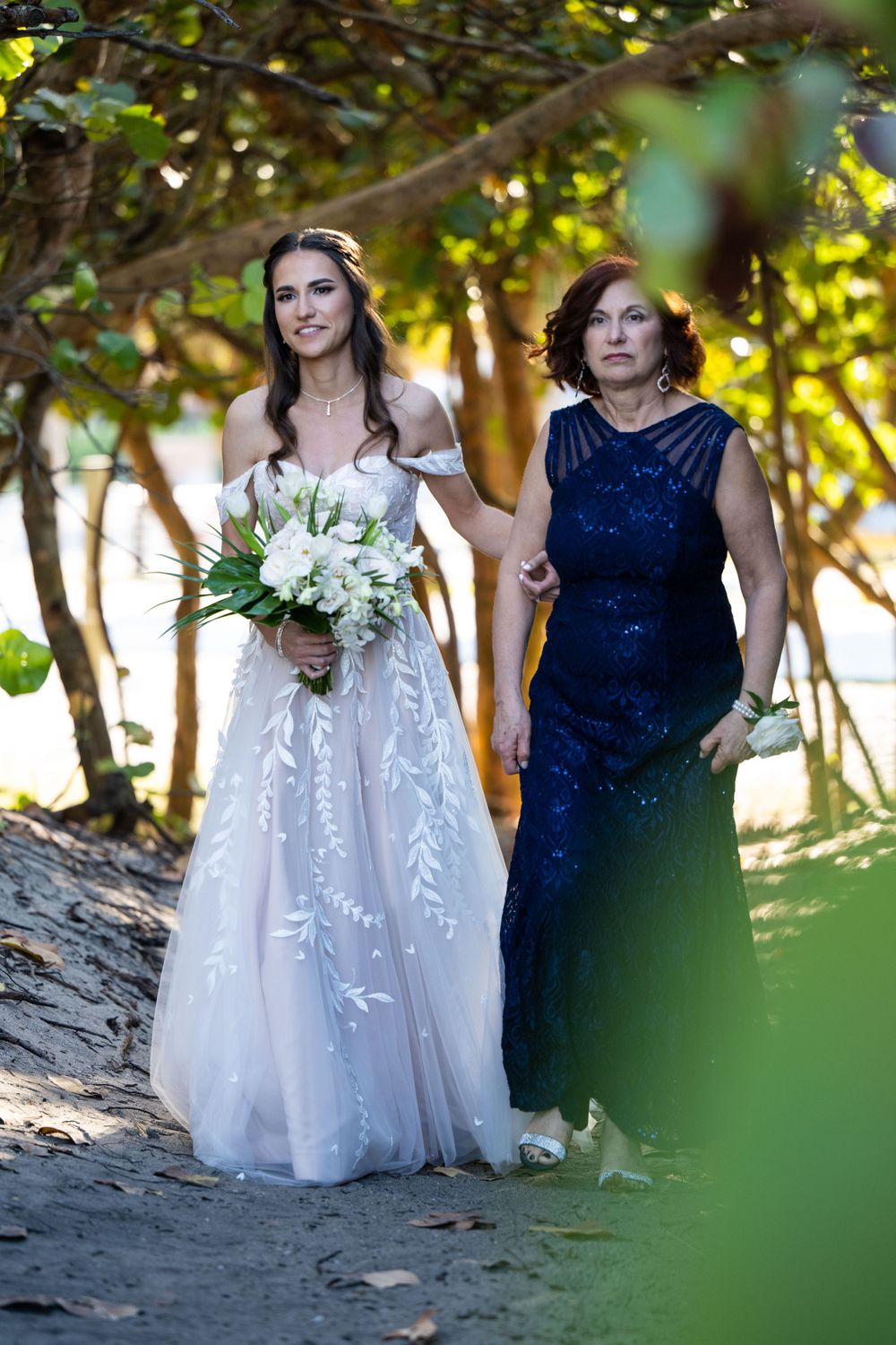 A bride in a white lace gown and companion in navy blue walk along a tree-lined path holding a white bouquet.