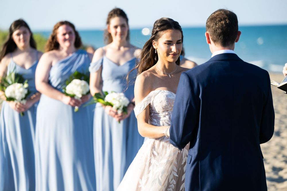 A beach wedding ceremony with bridesmaids in light blue dresses standing beside the couple on a sunny day.