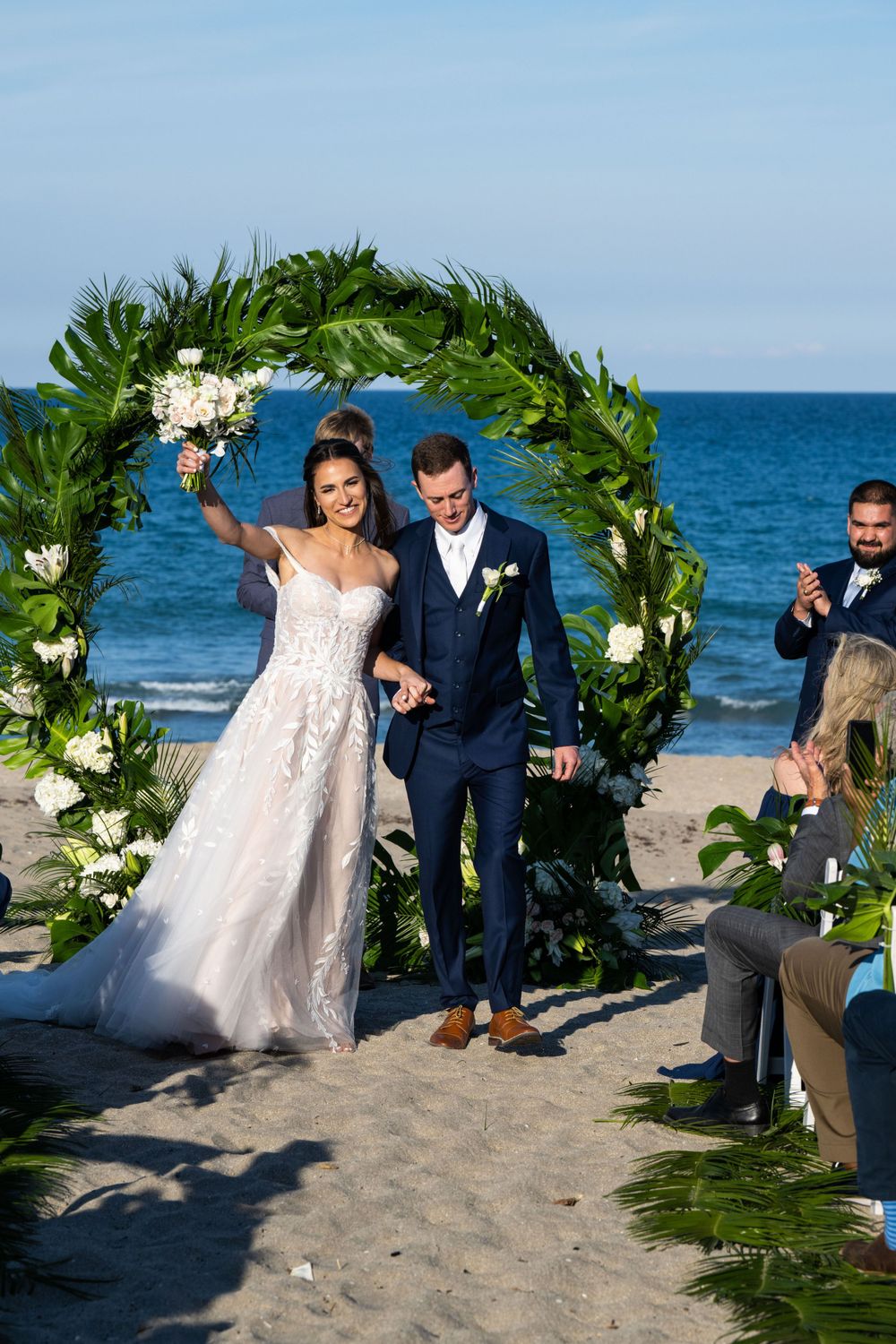 Newlyweds celebrate under a tropical floral arch on a beach with blue ocean backdrop.