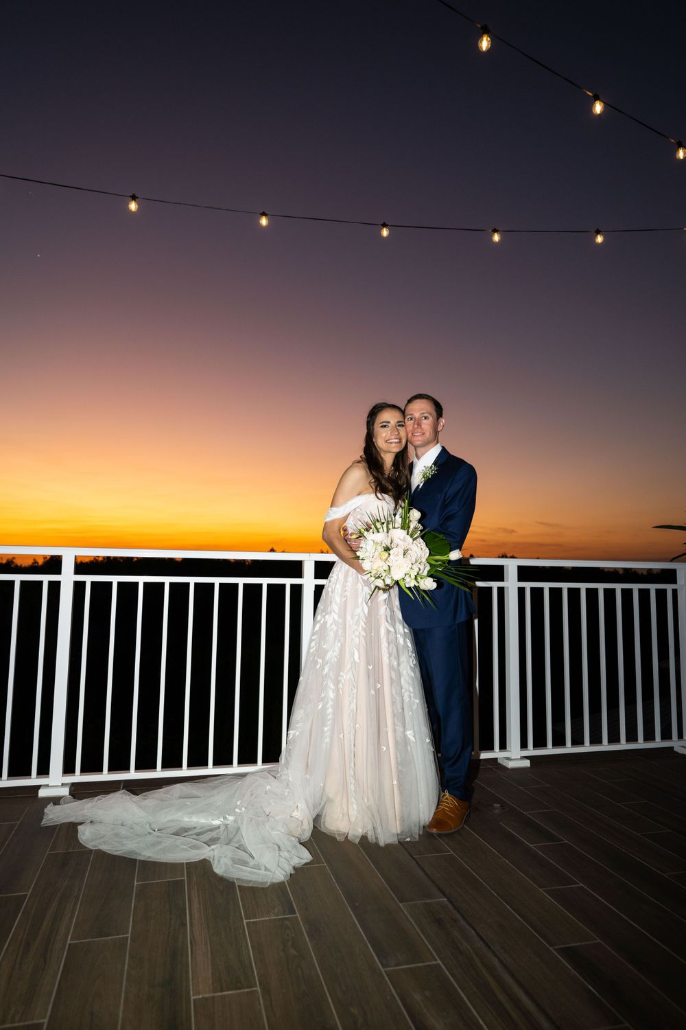 A couple shares an intimate wedding moment on a deck at sunset under string lights.
