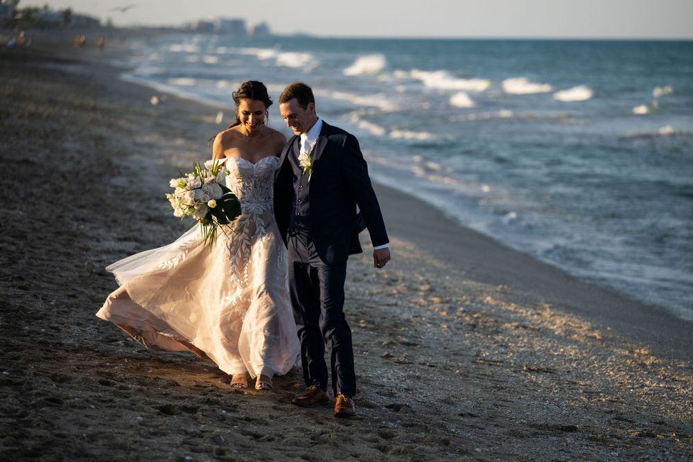 A couple walks along a beach shoreline with waves crashing and the bride's dress flowing in the wind.