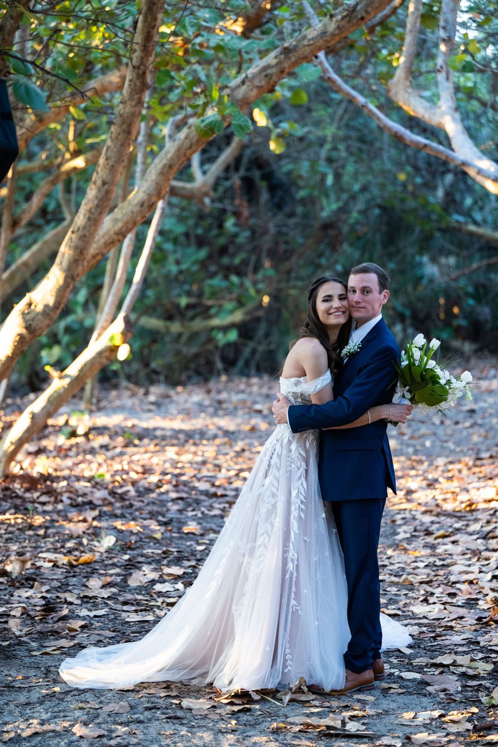 A romantic wedding couple embraces on a wooded path, with the bride wearing an off-shoulder lace gown.