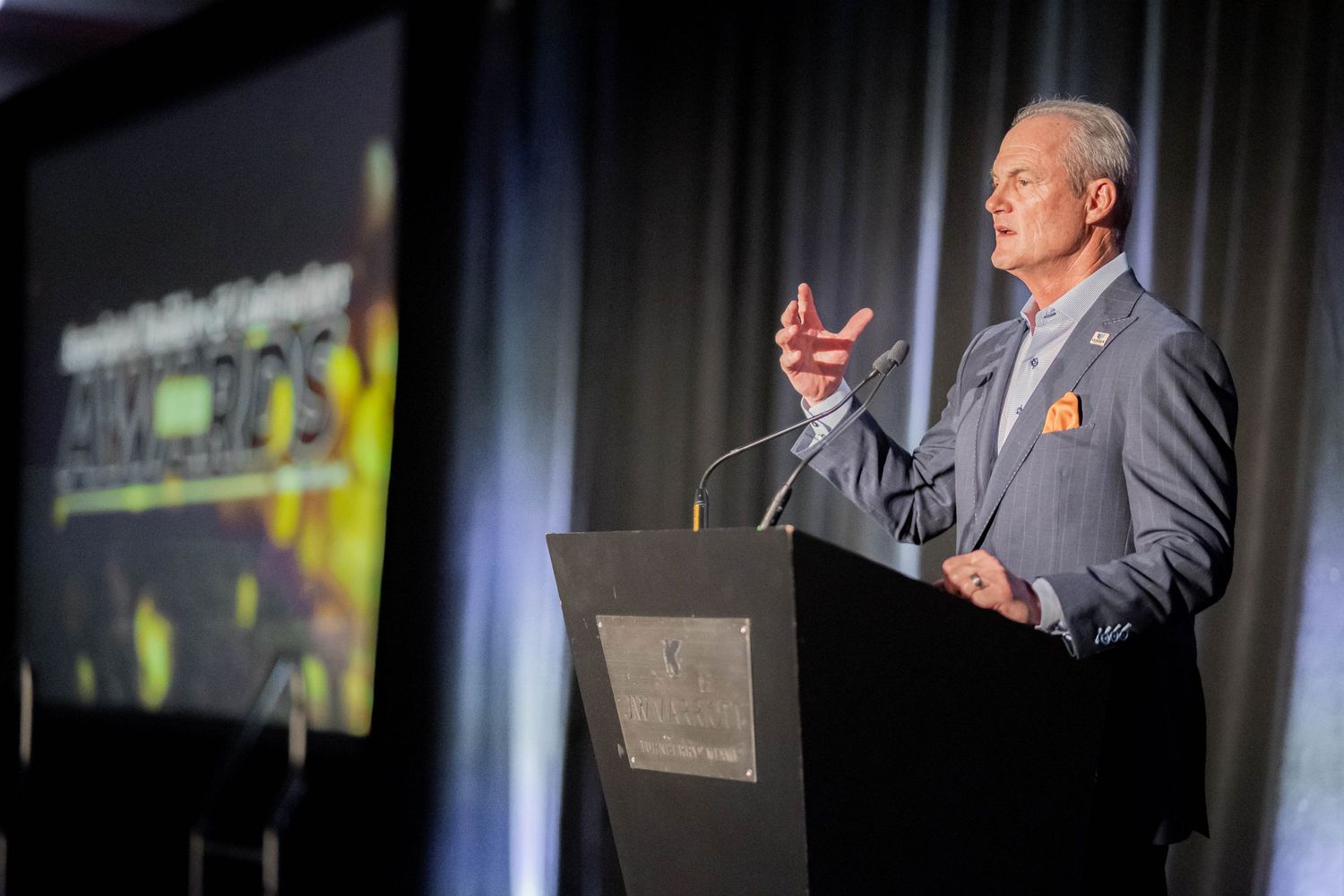 Professional speaker in gray suit delivers presentation at podium beside illuminated event signage on stage.
