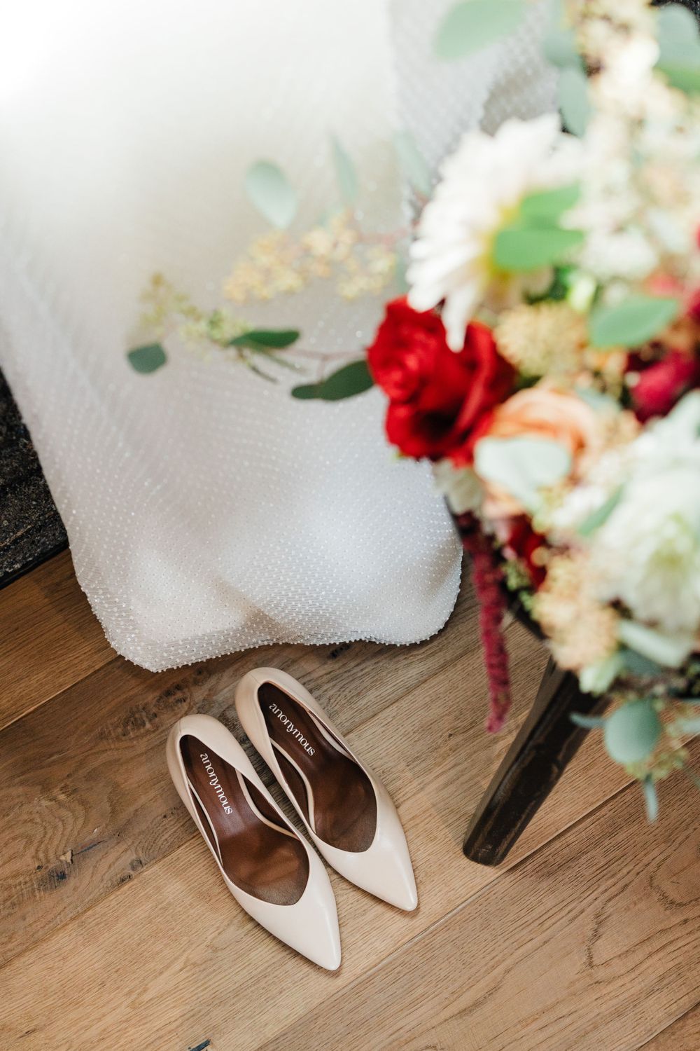 Elegant white pointed bridal shoes next to floral arrangement on wooden floor.