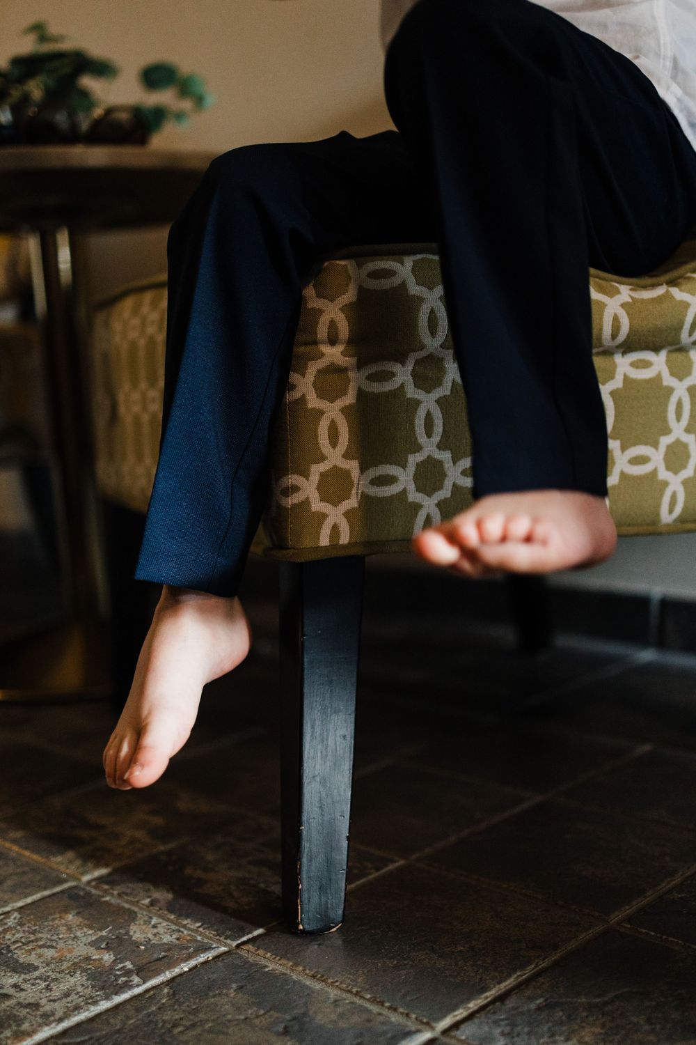 Bare feet dangling over a chair with patterned upholstery and dark wood flooring.