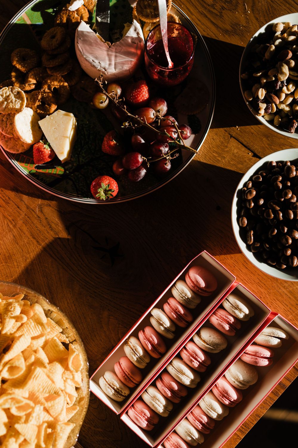 Elegant dessert spread with pink macarons, fresh fruits, and wine glasses on a wooden table in warm lighting.