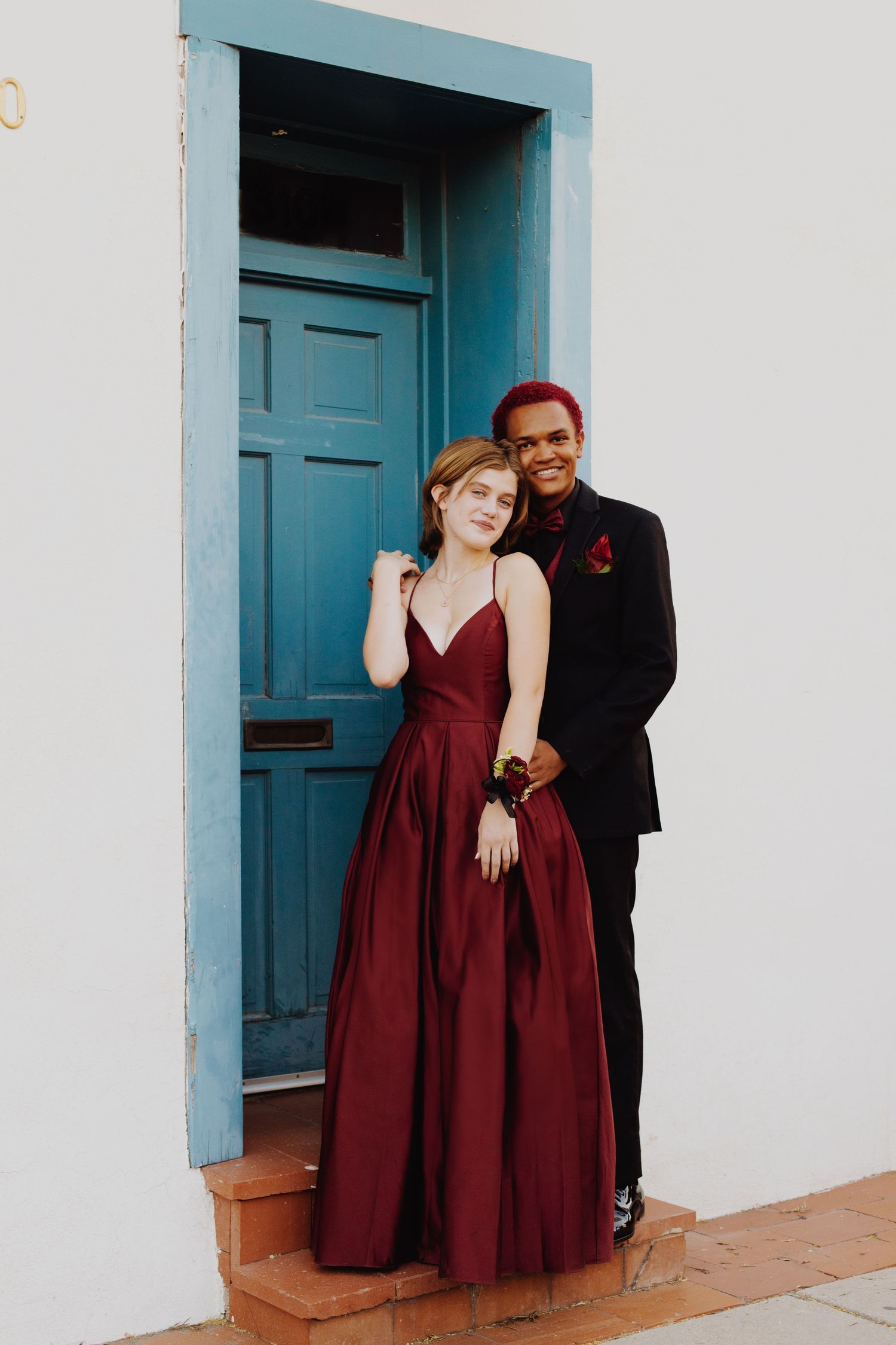 Couple in formal attire poses near turquoise doorway against white wall.