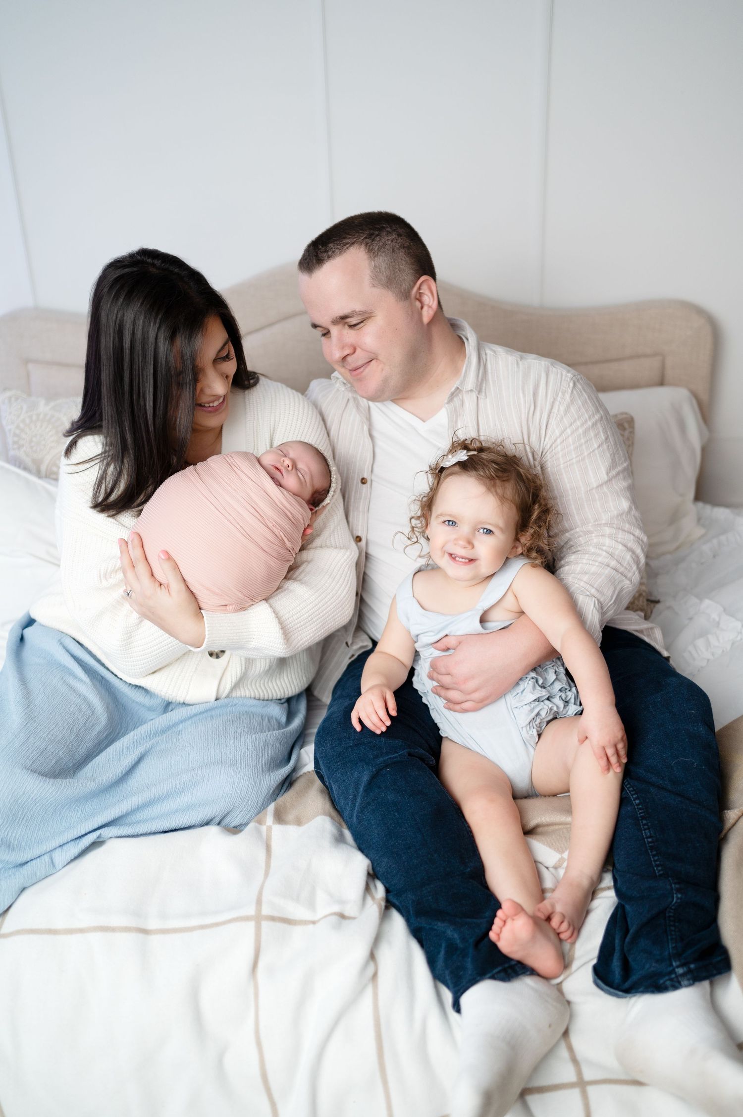 Intimate family portrait with parents gazing at newborn while toddler sits nearby.