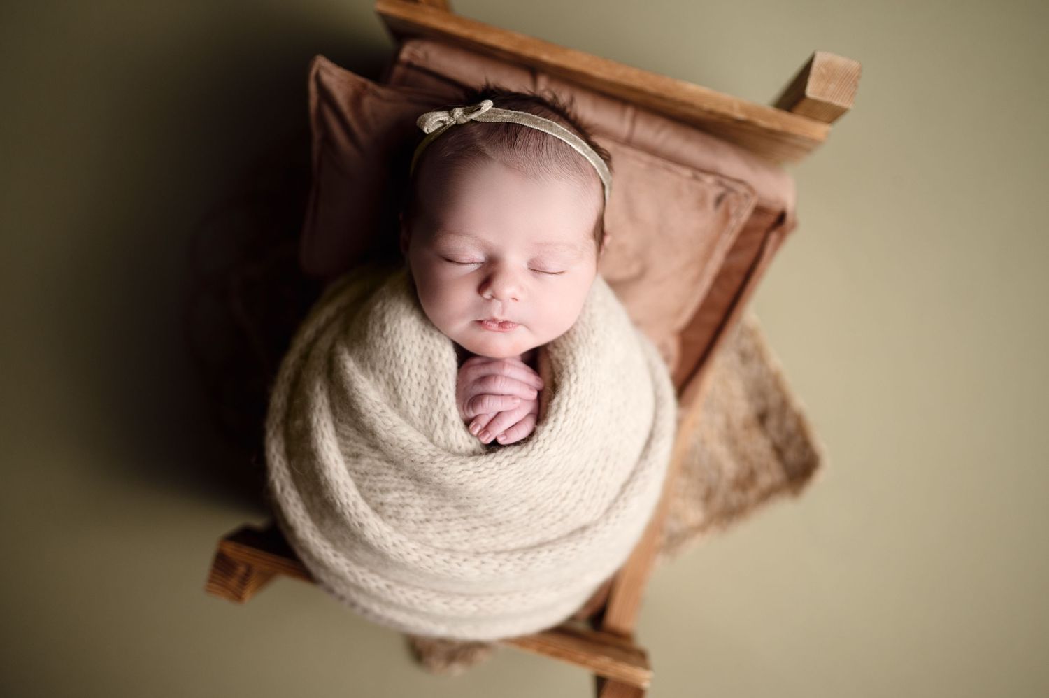 Sleeping newborn in cream wrap positioned on wooden prop with neutral background.