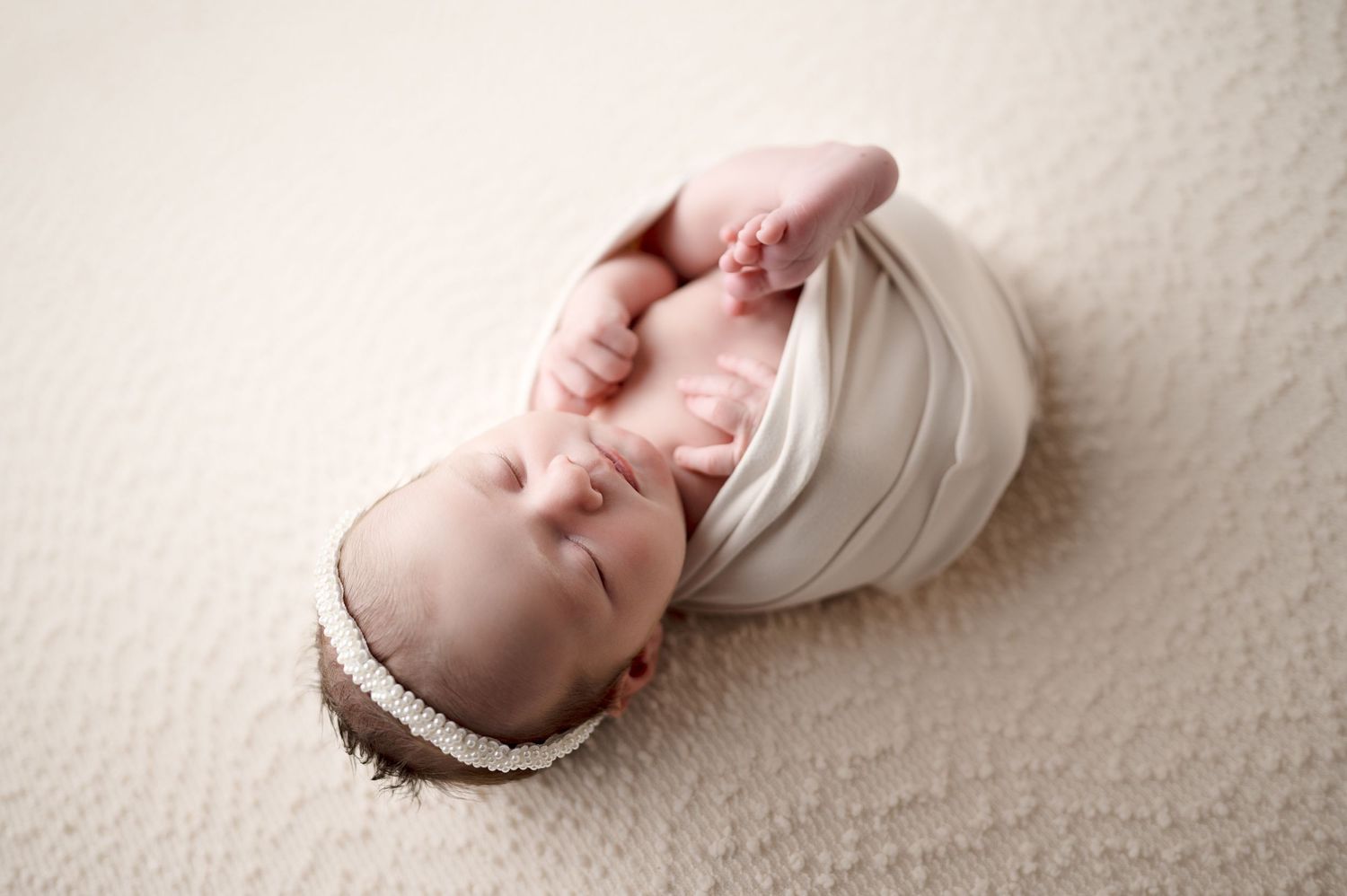 Newborn baby wearing white headband sleeping peacefully on cream backdrop.