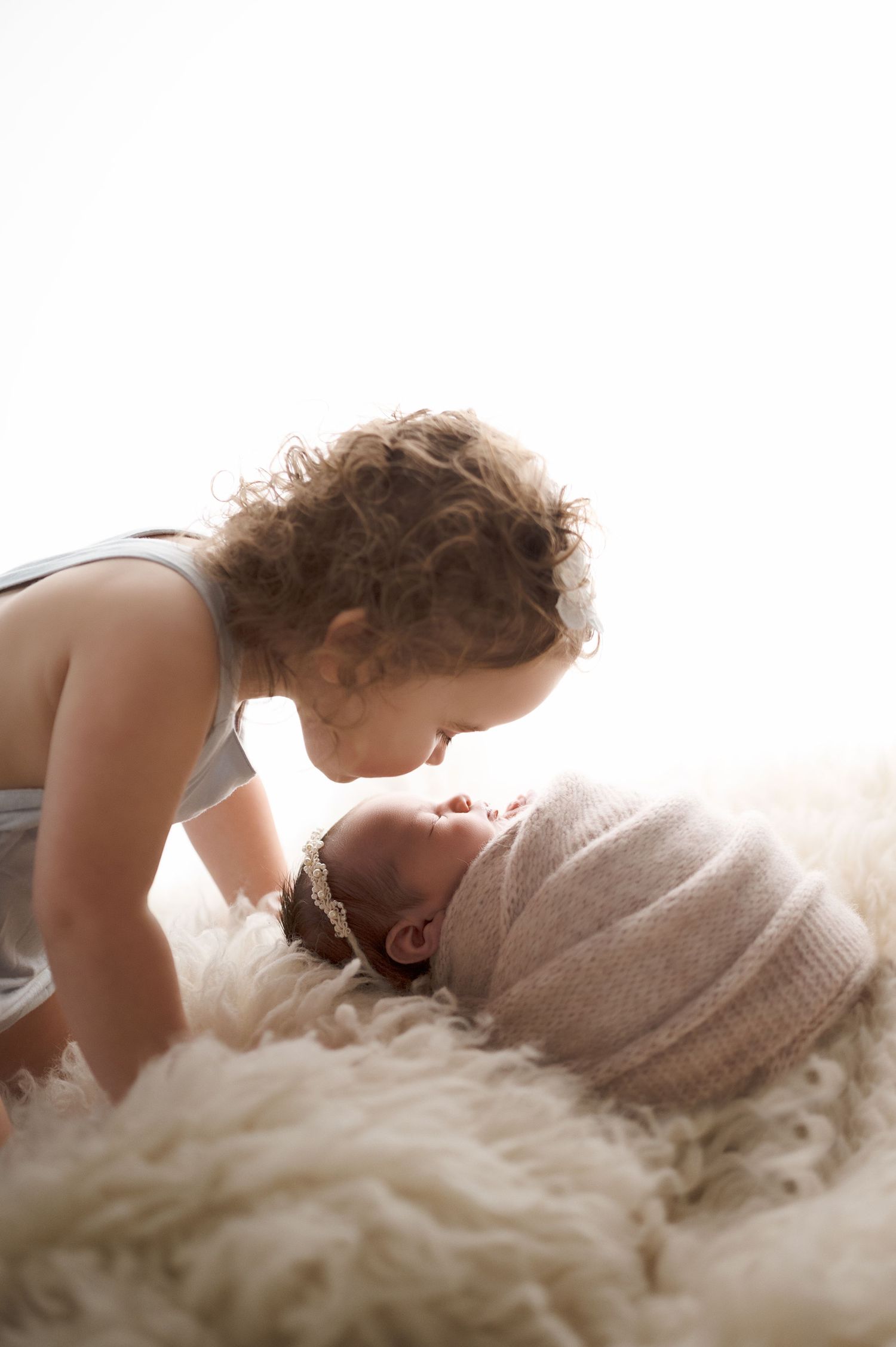 Toddler in gray outfit leans over to kiss sleeping newborn on fluffy white blanket.
