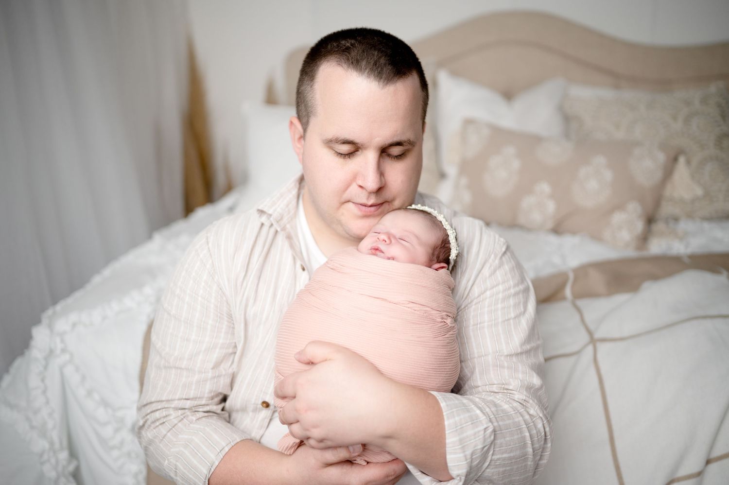 Father in light sweater holding sleeping newborn wrapped in pink swaddle.