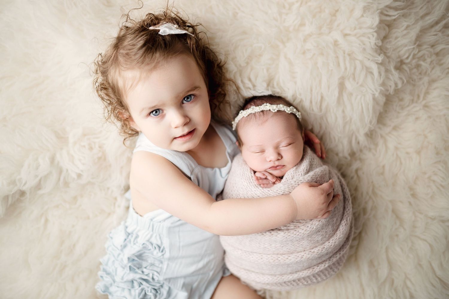 Sweet sibling embrace on white fur as older child holds swaddled newborn.