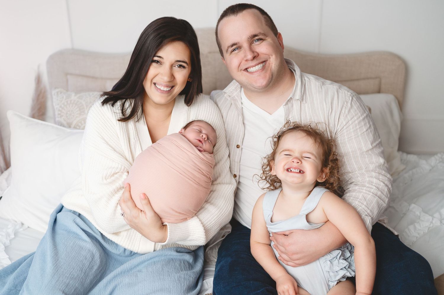 Happy family poses together on bed with newborn wrapped in pink and toddler smiling.
