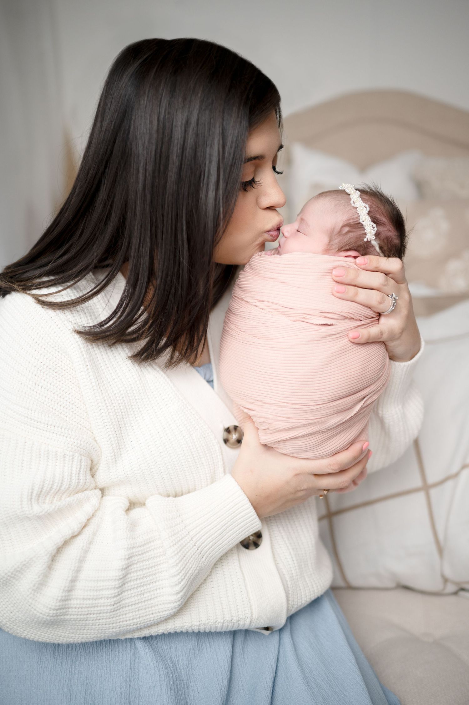 Mother in white cardigan kissing swaddled newborn baby on bed.