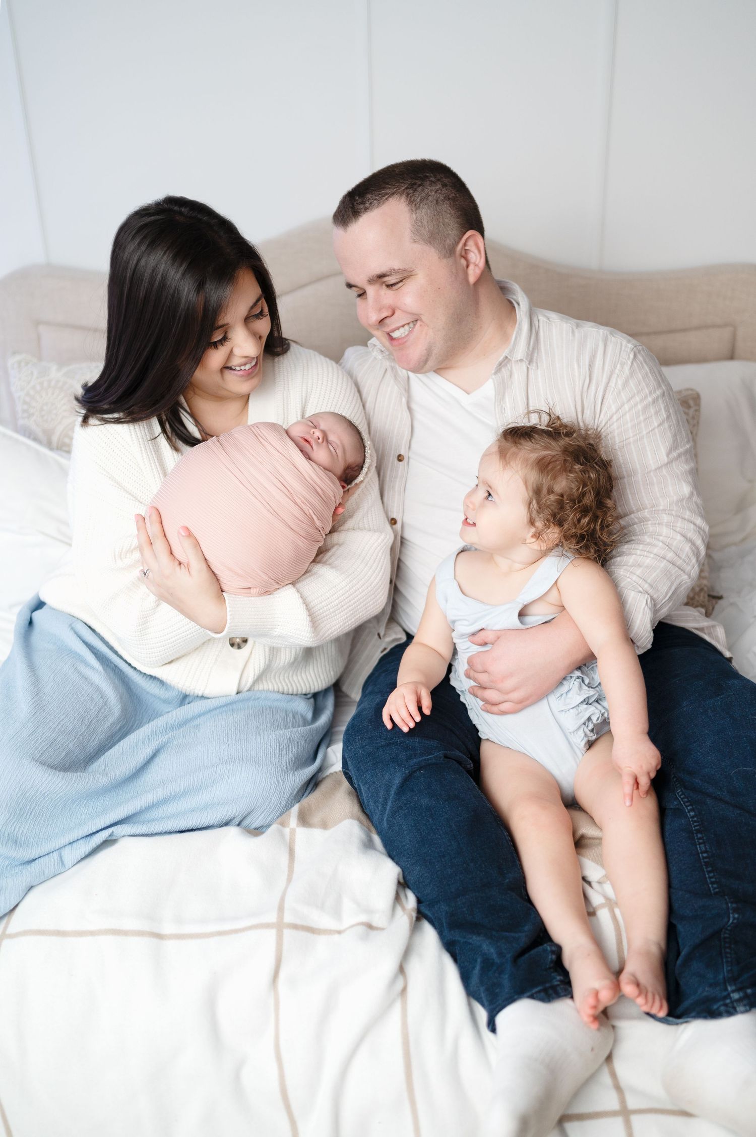Candid family photo showing parents with newborn and toddler relaxing on bed.