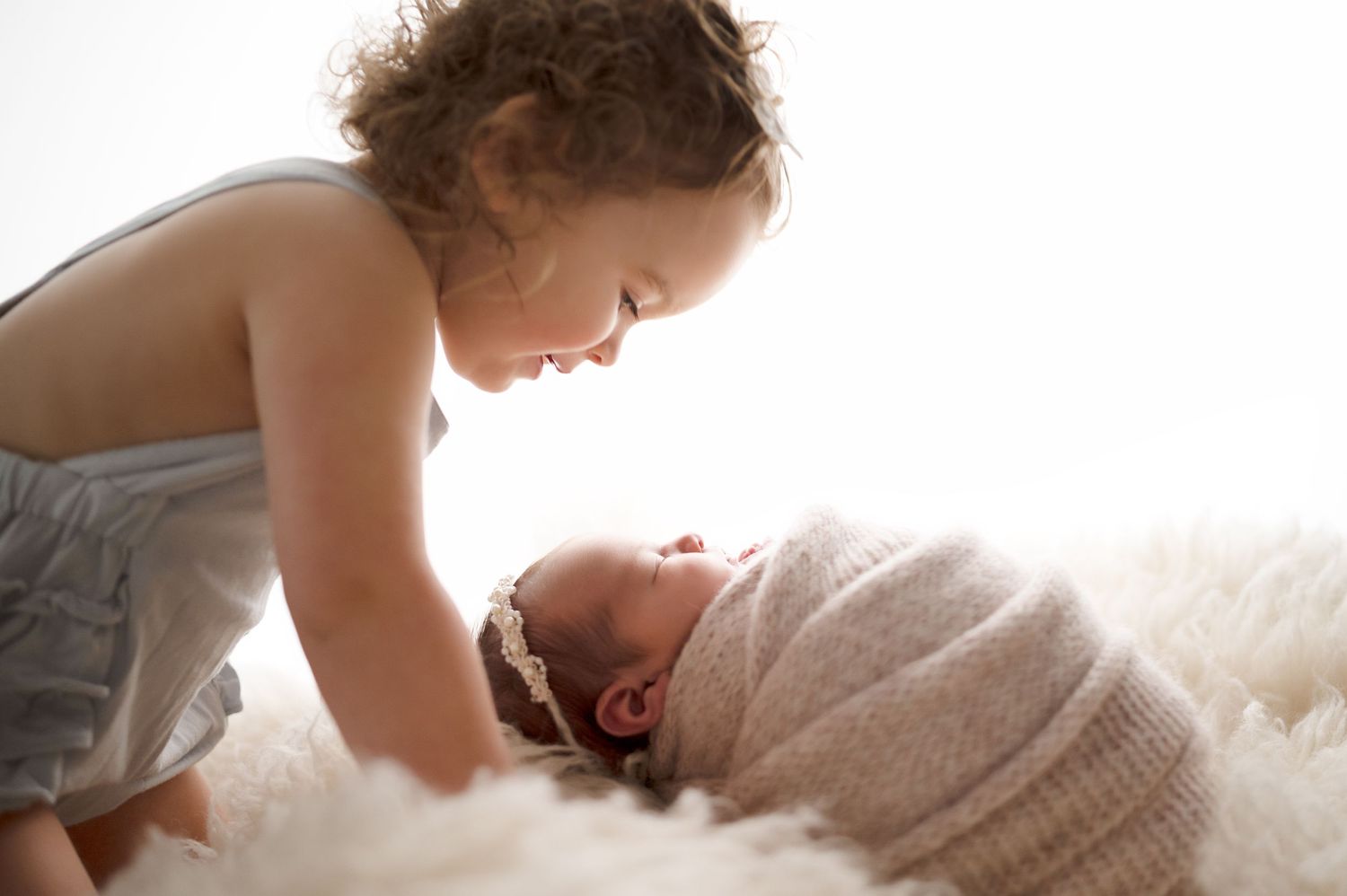 Tender sibling moment captured as older child leans over wrapped newborn on white fur.