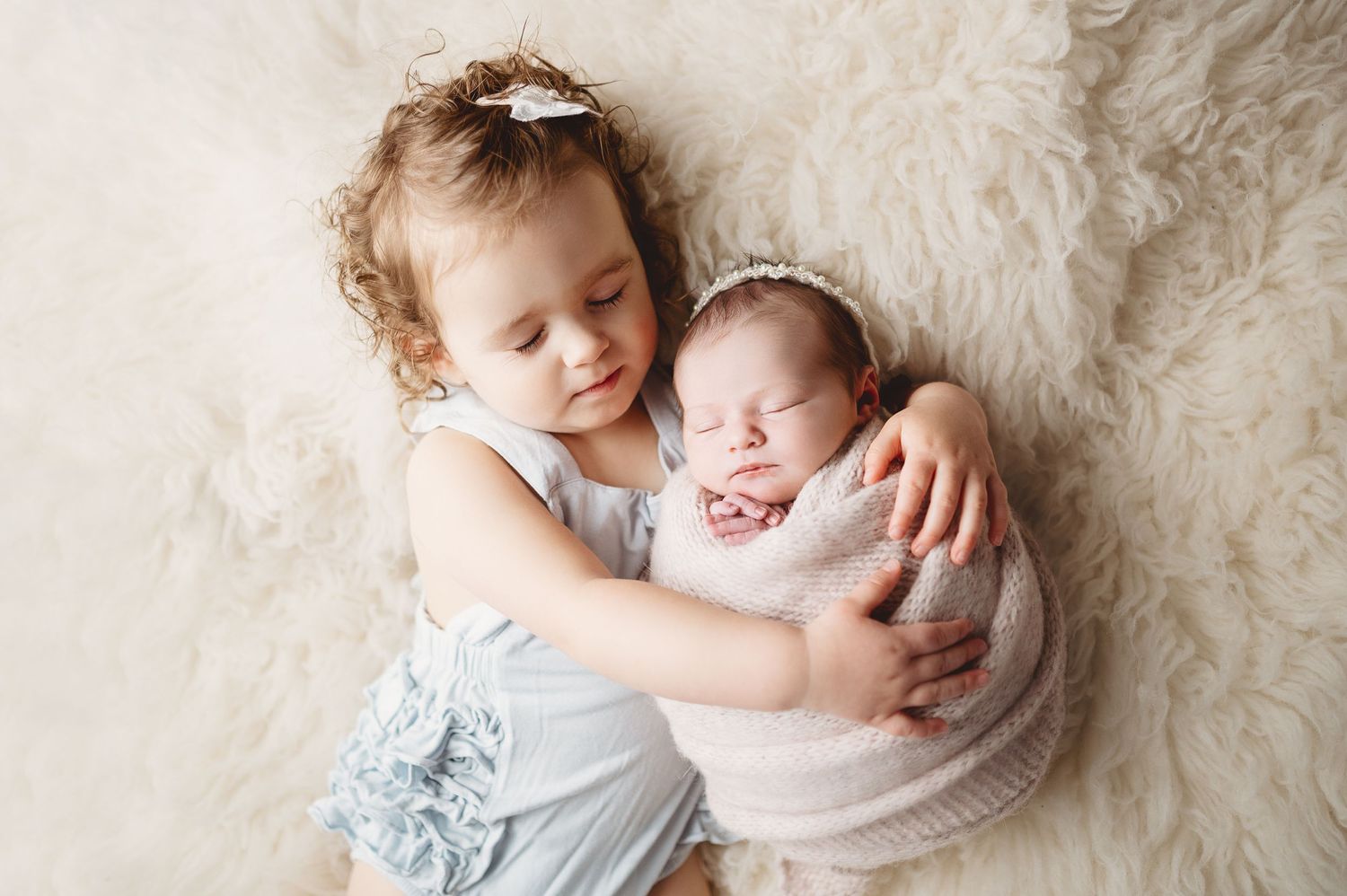 Joyful sibling moment as child in blue dress cuddles wrapped newborn on white fur.