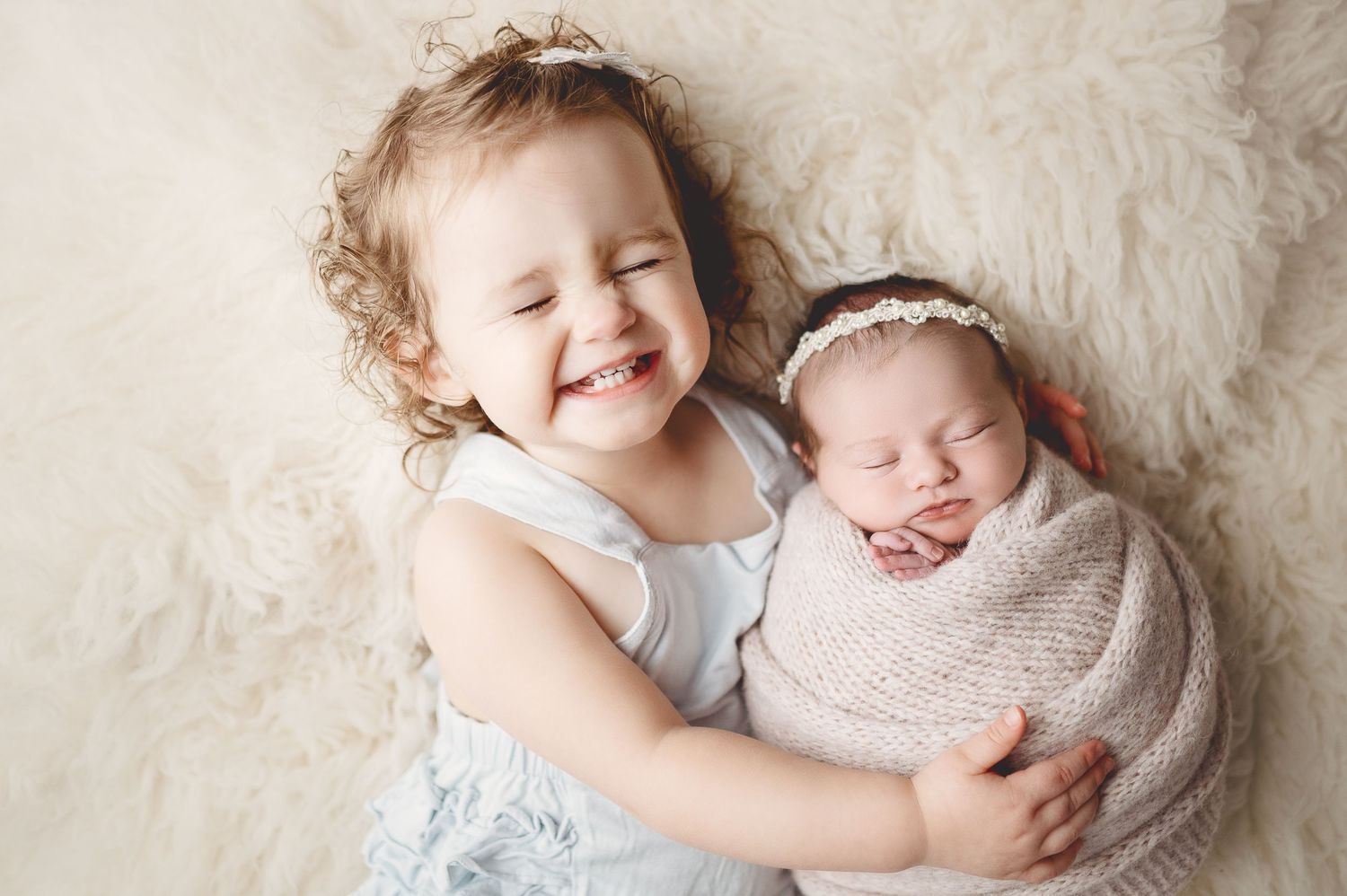 Two siblings share laughter and cuddles on soft white fur background.
