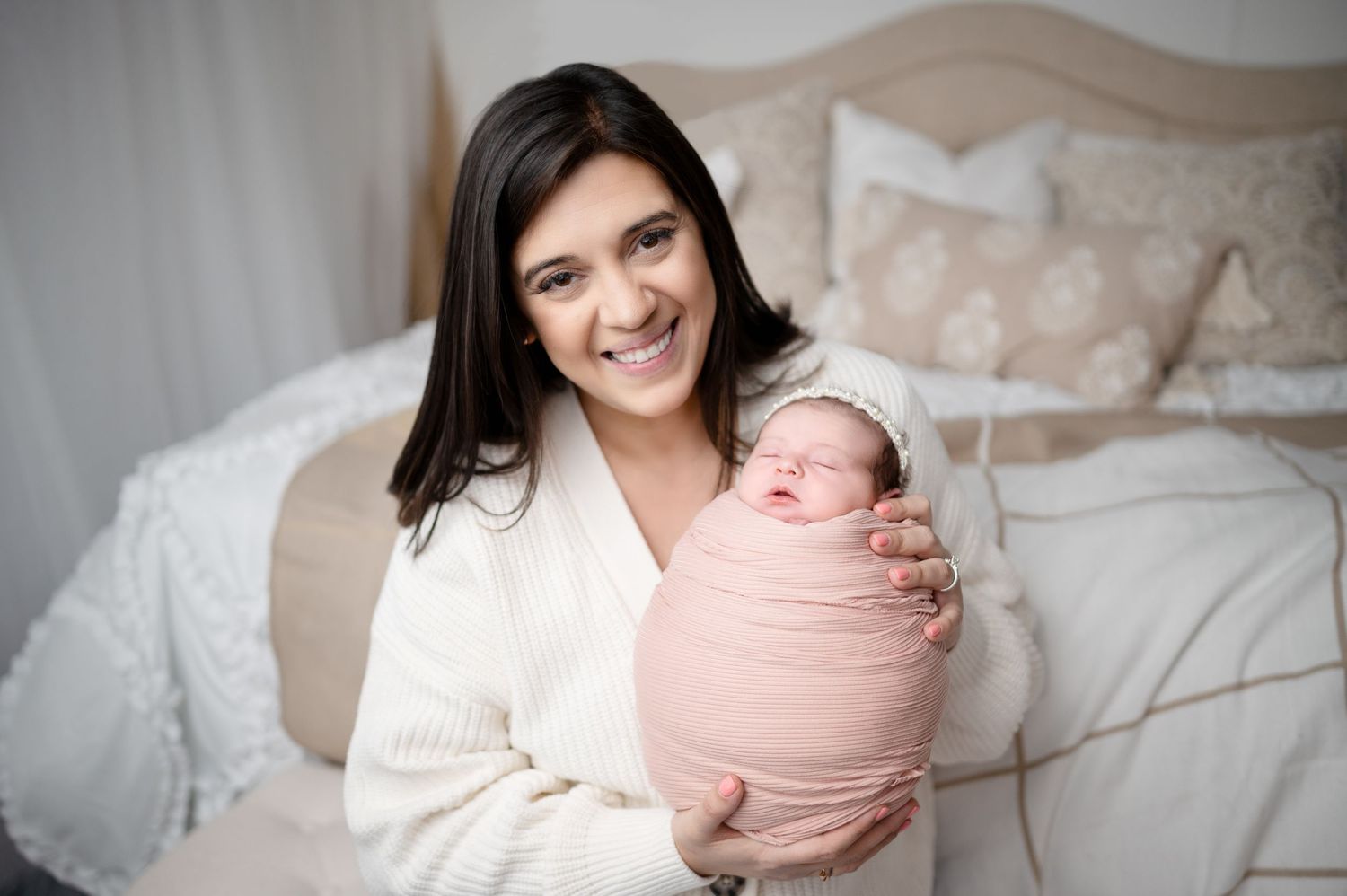 Tender moment between mother in white sweater holding swaddled newborn on bed.