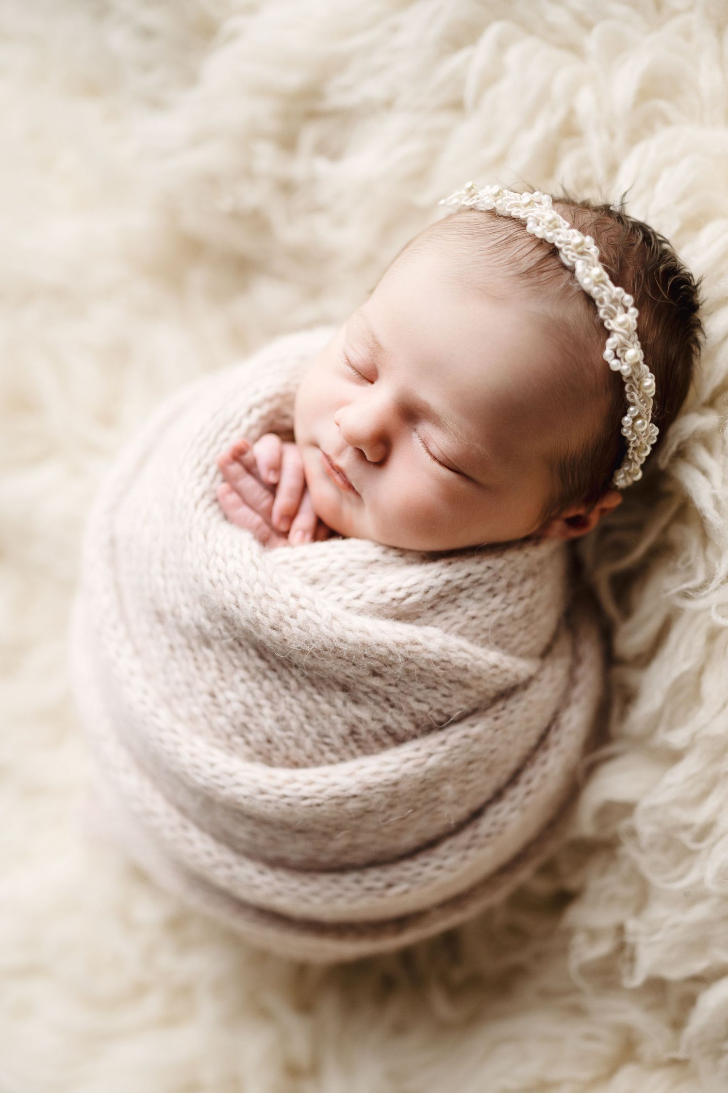 Sleeping newborn in cream knit wrap with delicate lace headband on fluffy white backdrop.