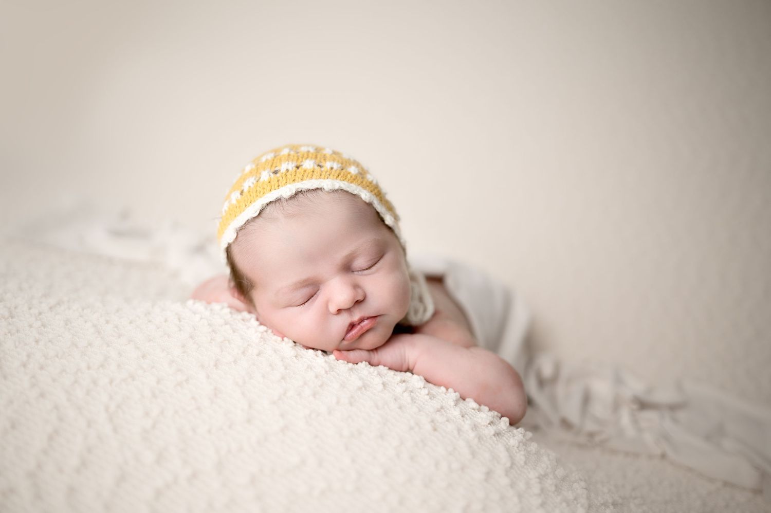 Peaceful newborn baby wearing yellow crochet hat sleeping on white textured surface.