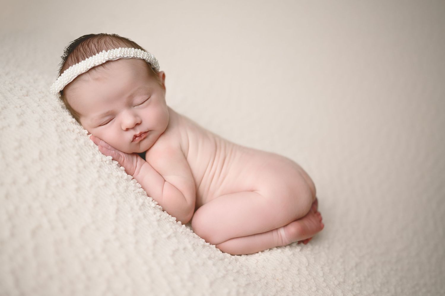Sleeping newborn with delicate headband rests peacefully on textured cream blanket.