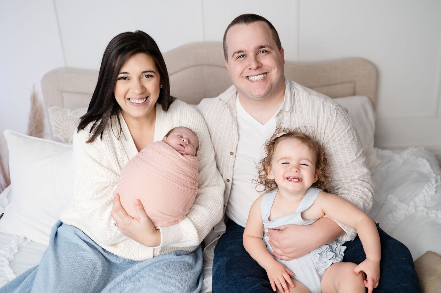 Family portrait with newborn and toddler sitting together on white bedding.