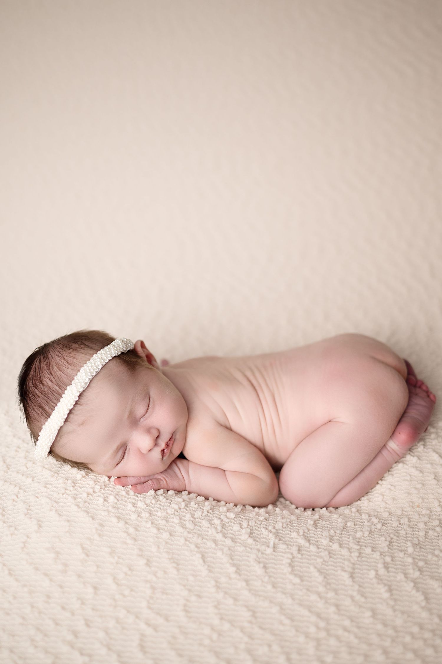 Sleeping newborn in white headband curled up on textured cream blanket.