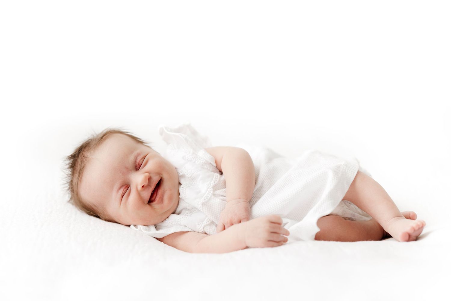 A sleeping baby wearing white clothing smiles while lying on soft white bedding.
