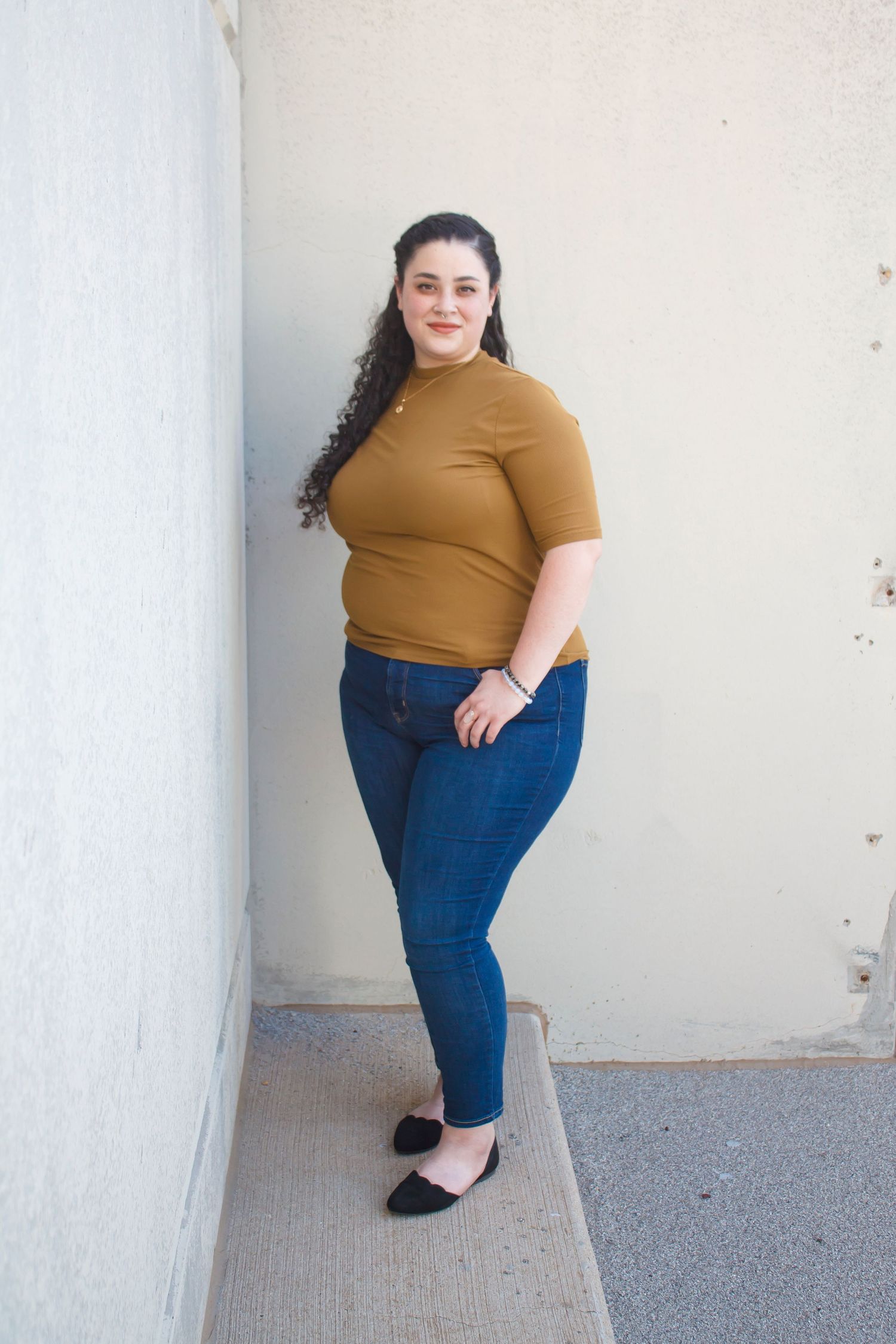 Full length portrait of someone in a mustard top and jeans against a light wall.