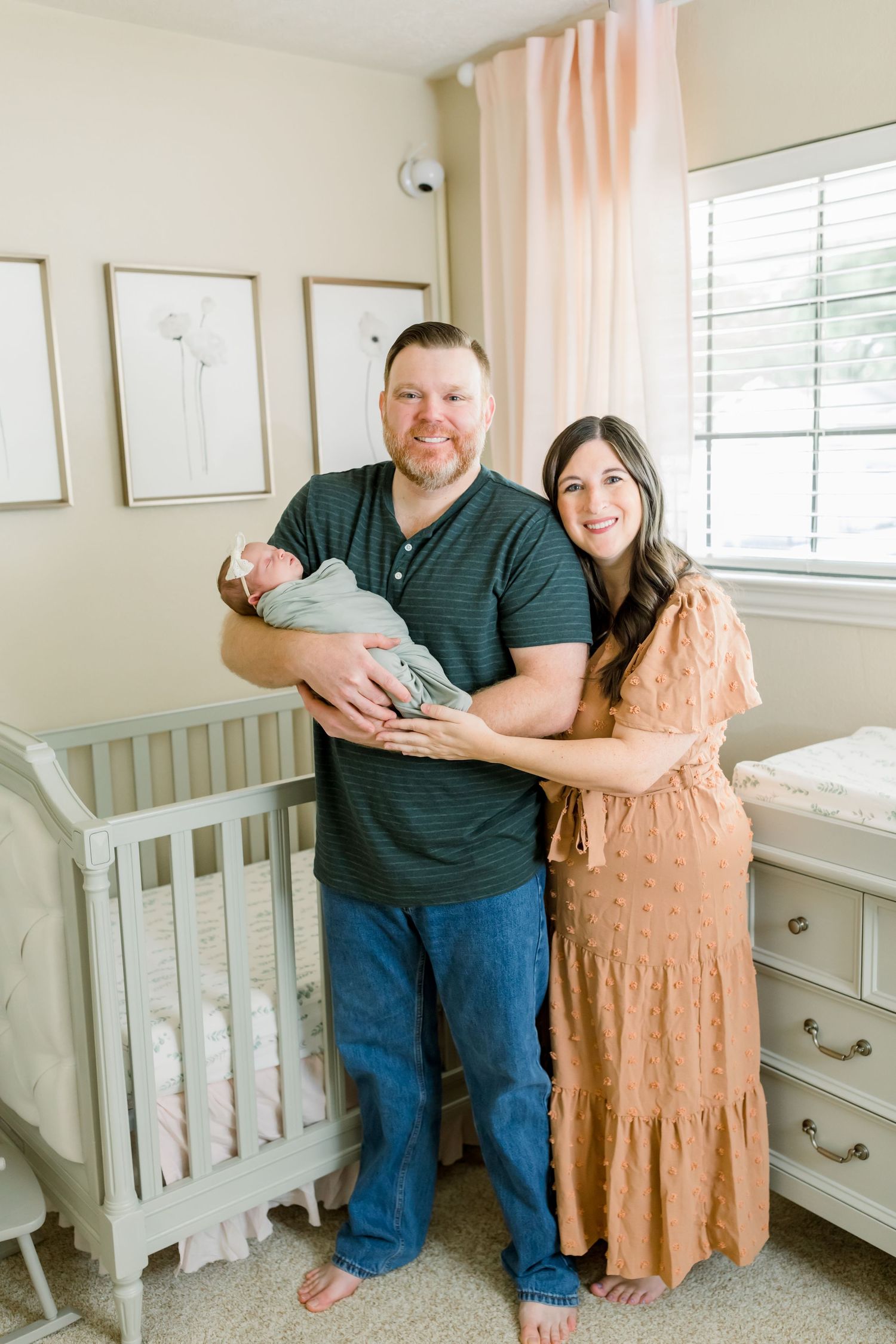 A couple stands together holding their newborn baby in a serene nursery with neutral decor.