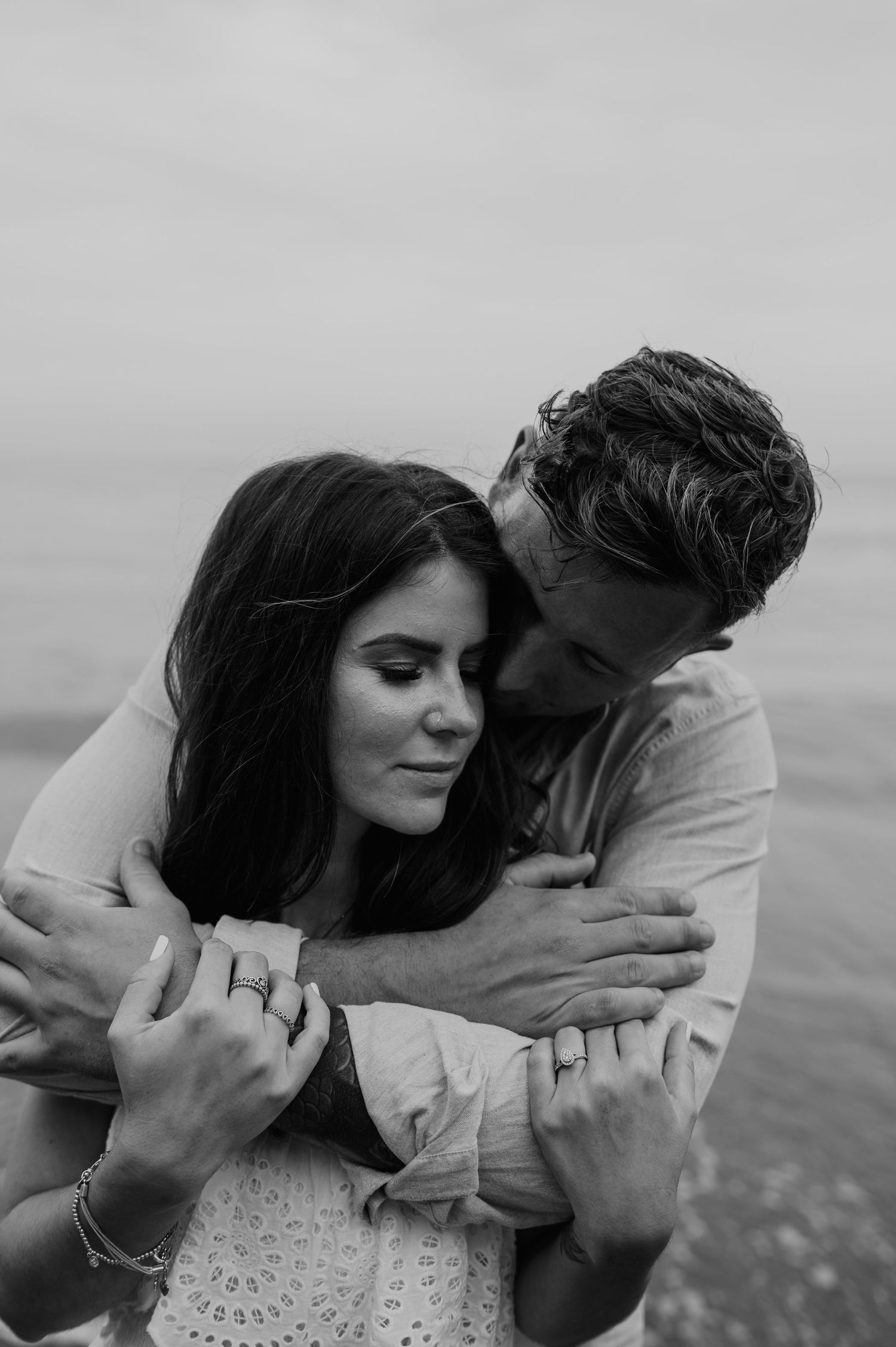 Romantic black and white photo of embracing couple by the water with engagement ring visible.