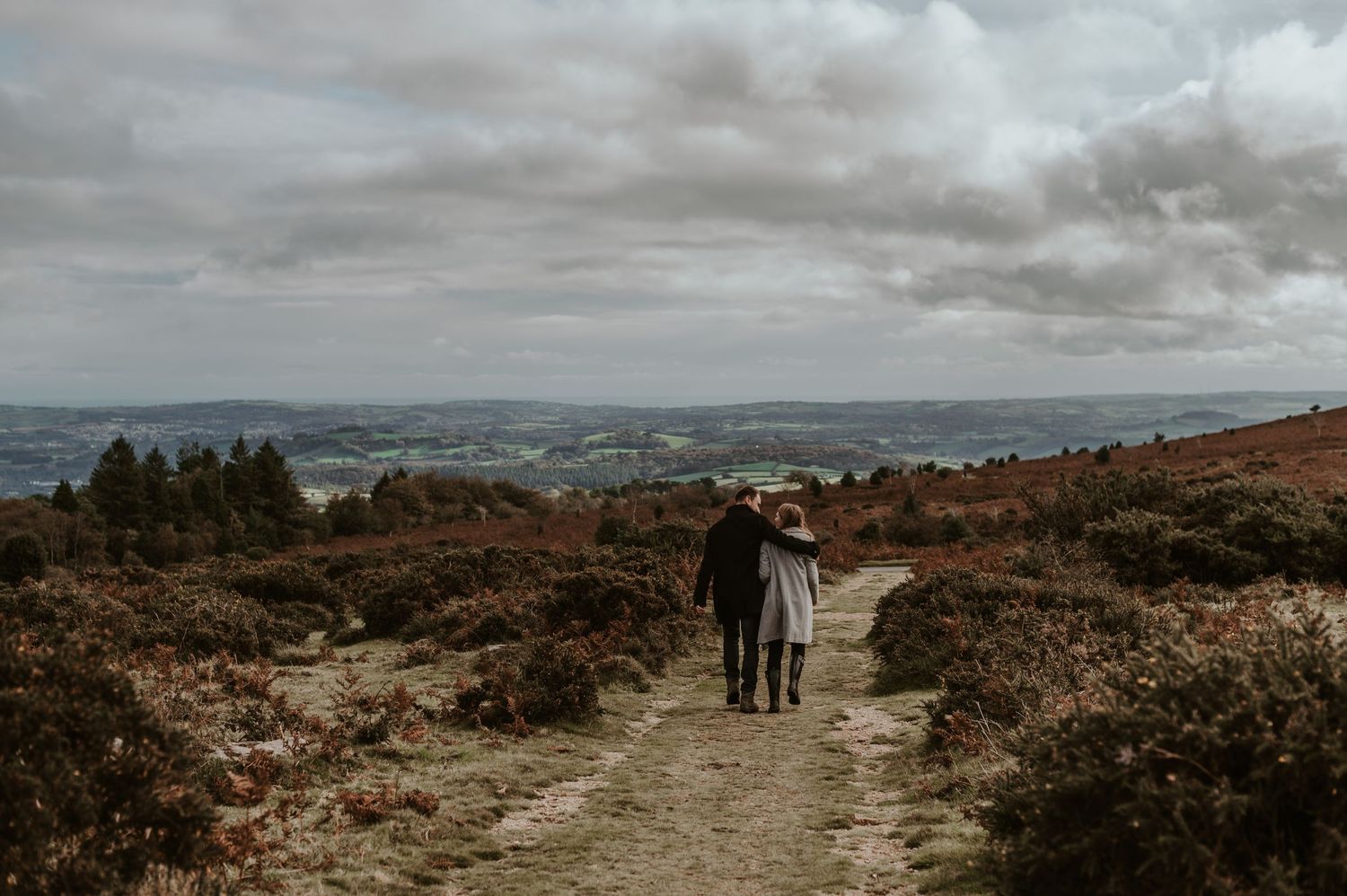 A couple walks along a winding hiking trail through heathland on a cloudy day with dramatic skies and distant views on Dartmoor.